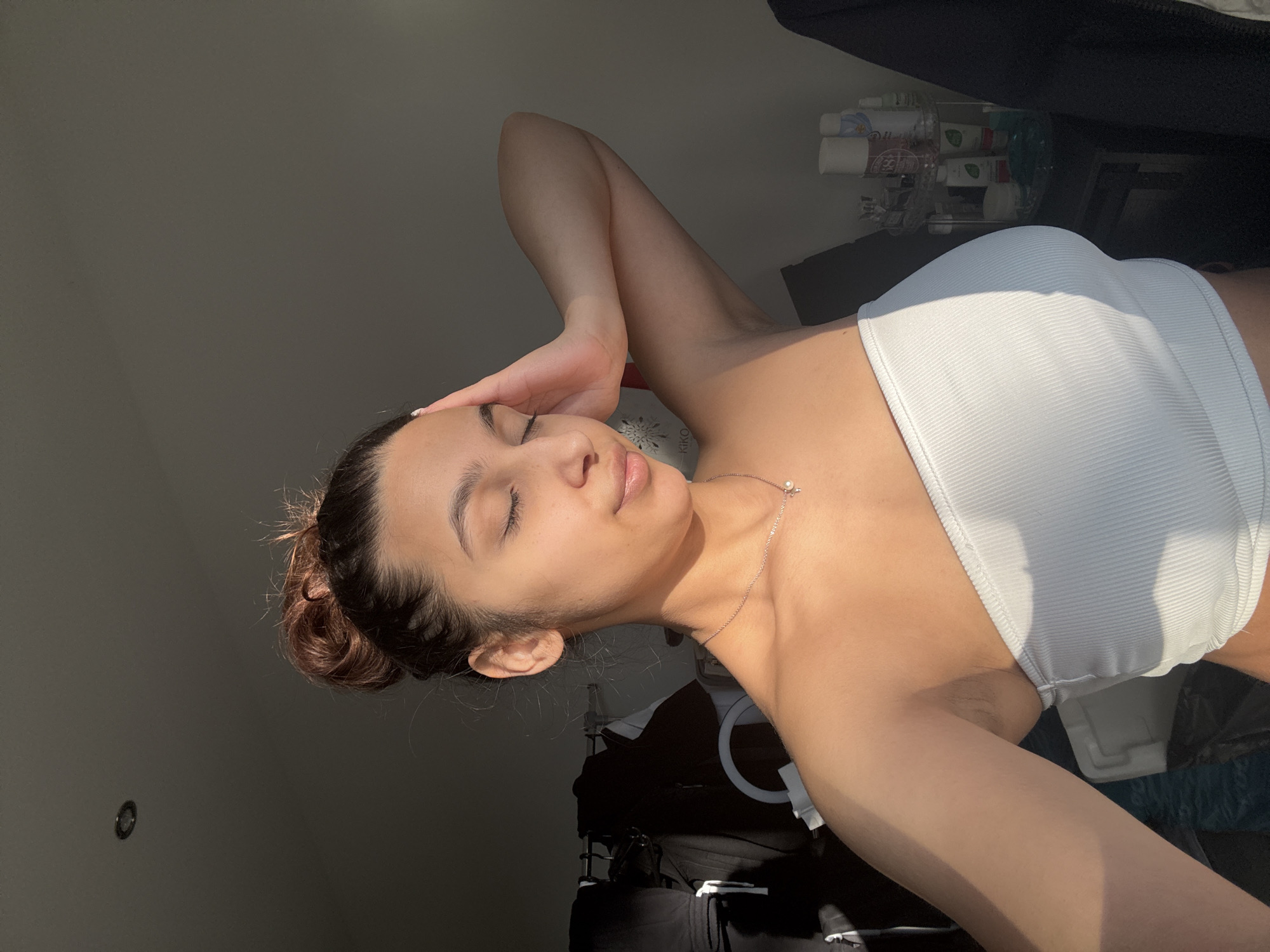 A woman is posing indoors in Mönchengladbach, Germany. She is wearing a white tube top and a delicate necklace. Her eyes are closed and her hand is near her forehead. Behind her are various objects and furniture. The lighting is uneven with some areas brightly lit by sunlight.