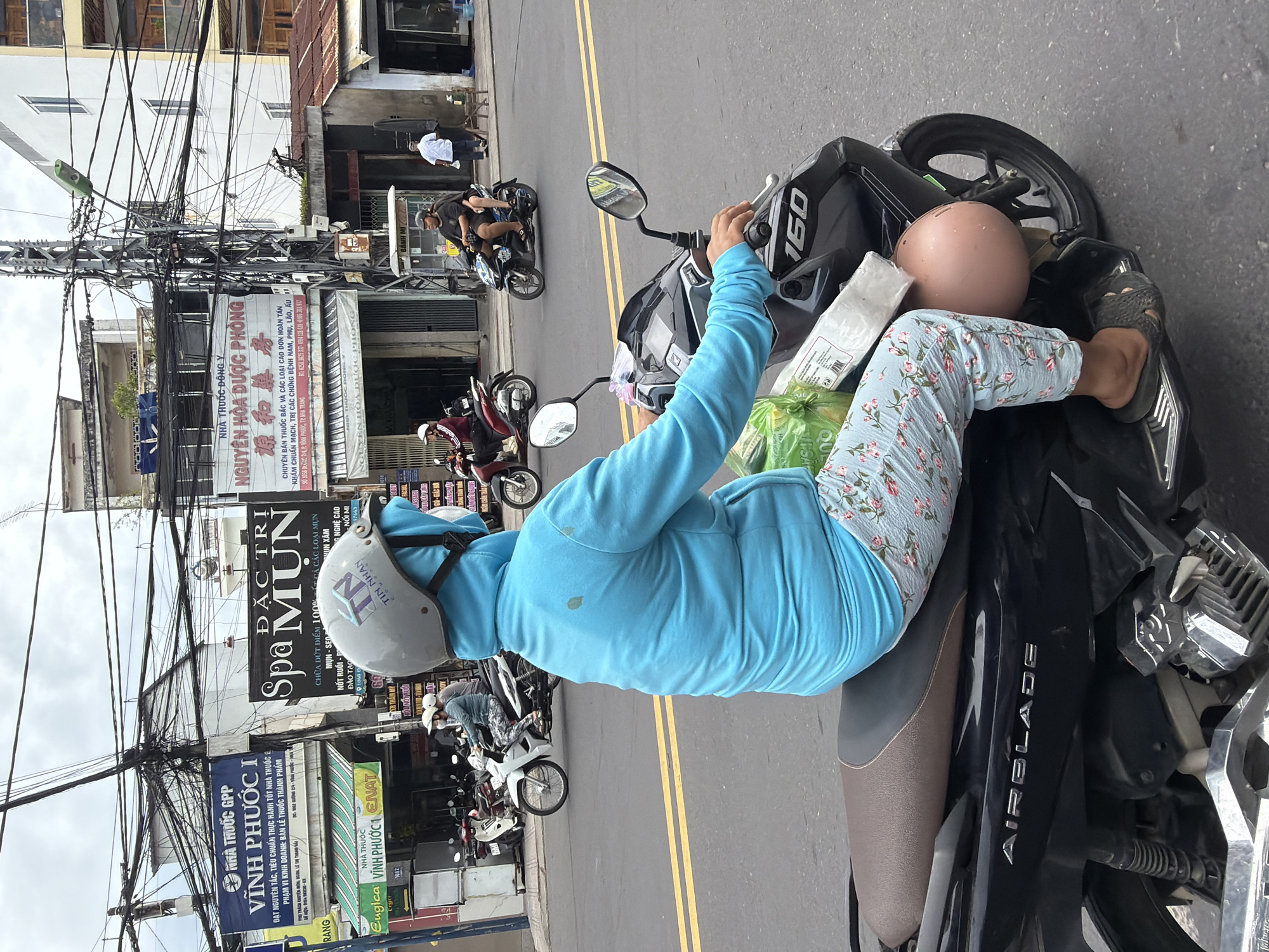 In Nha Trang, Vietnam, a person wearing a blue long-sleeved shirt and floral patterned pants rides a black Honda Airblade 160 motorcycle. They are wearing a light grey helmet with a blue face covering and carrying a plastic bag with green items inside. The rider's left hand is on the handlebar, and their right foot is on the footboard. The motorcycle has a pink helmet hanging from the front. In the background, buildings with various signs are visible, including "VINH PHUOC 1 ENAT," "Spa MUN," and "NGUYEN HOA DUOC PHONG." Other motorcycles are parked and in motion, and utility wires are strung across the sky. The scene appears to be on a city street with a yellow dividing line on the asphalt road.