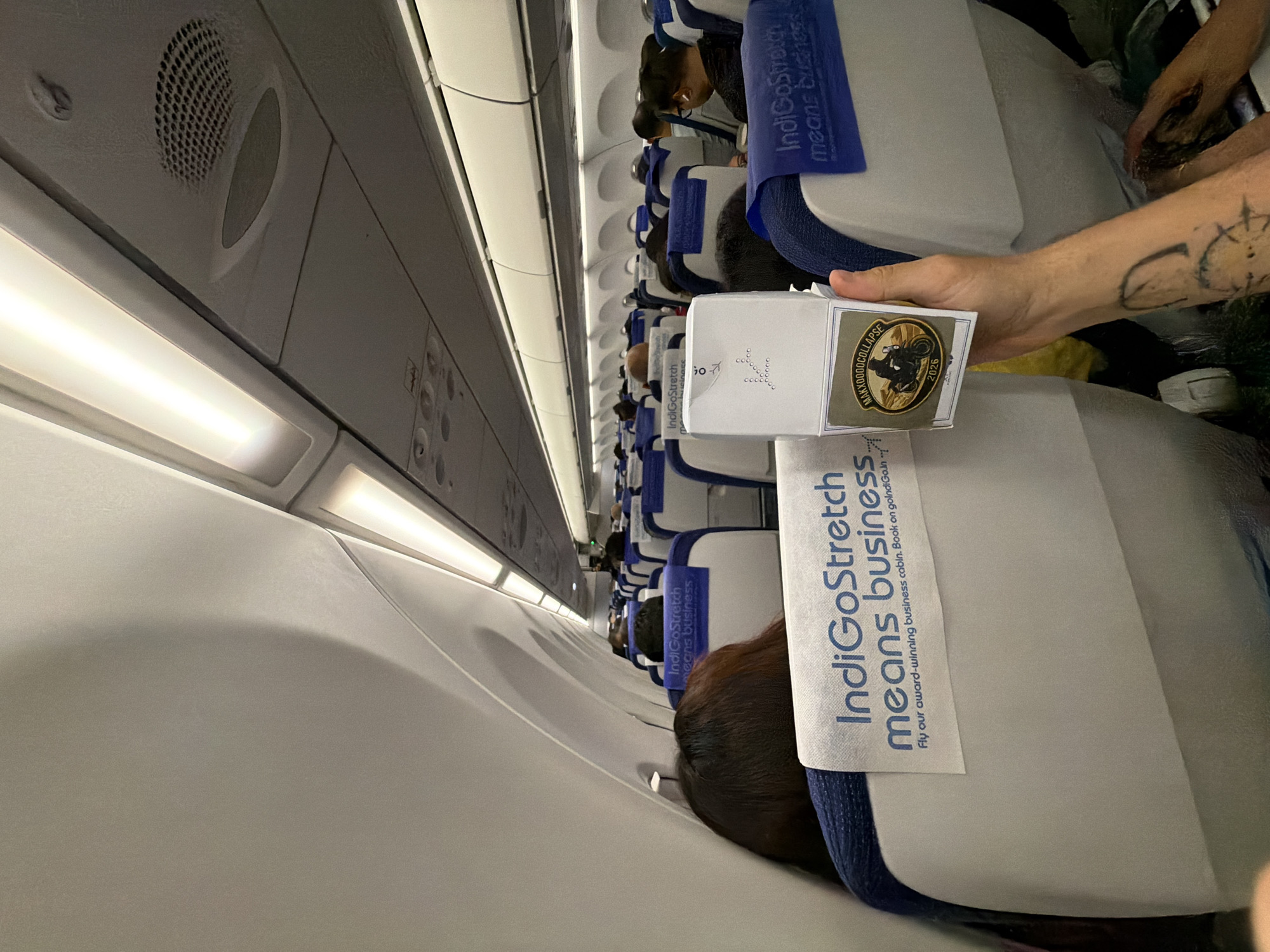 The image shows the interior of an airplane cabin, likely during a flight, as indicated by the rows of seats and overhead compartments. The seats are upholstered in blue and white, and headrest covers display the IndigoStretch logo and the tagline "means business." In the foreground, a person's arm, adorned with a tattoo, is holding a small, white rectangular object with a label that features an image of a person riding a motorcycle and the year "2026." The lighting appears to be artificial, emanating from the overhead strip lights. The scene is viewed from the perspective of a passenger seated on the left side of the plane, looking towards the front. While the image depicts an airplane interior, there is no information within the image itself to suggest it is located in Nadergul, India.