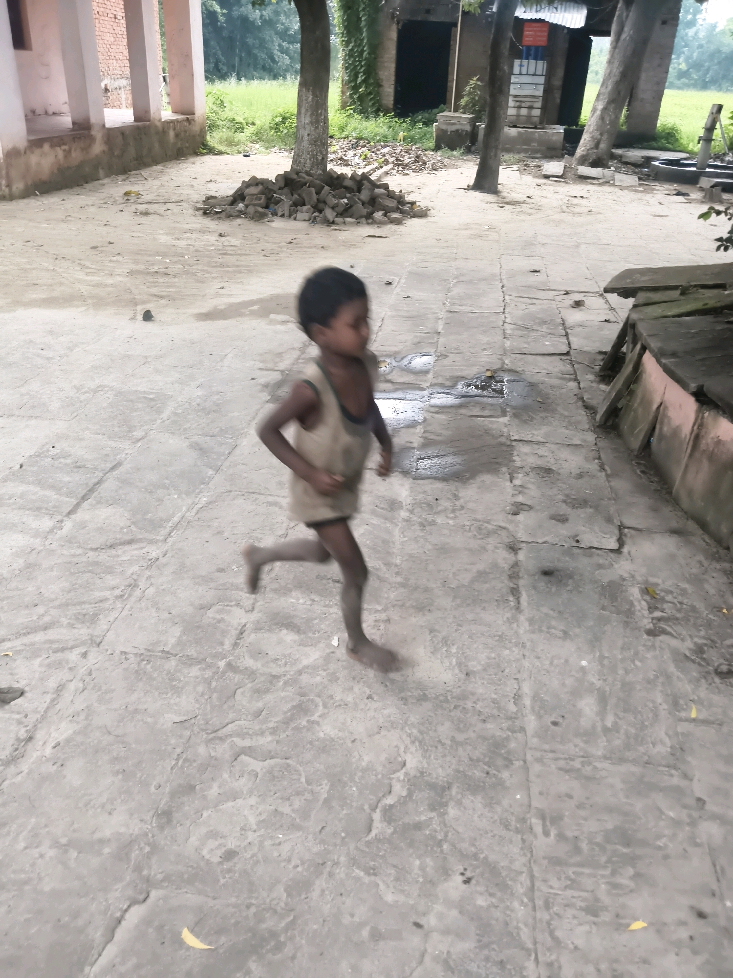 A young boy, wearing a sleeveless undershirt and shorts, is captured in motion, running across a paved courtyard in Rajbaliya, India. The boy's limbs are blurred due to the speed of his movement, suggesting a dynamic action. The ground is composed of large, rectangular stone slabs, some of which appear wet or stained. Scattered debris, including bricks and fallen leaves, is visible on the ground. In the background, a building with pillars and a brick wall is present, along with trees and foliage. The overall environment appears to be an outdoor, semi-urban or rural setting.