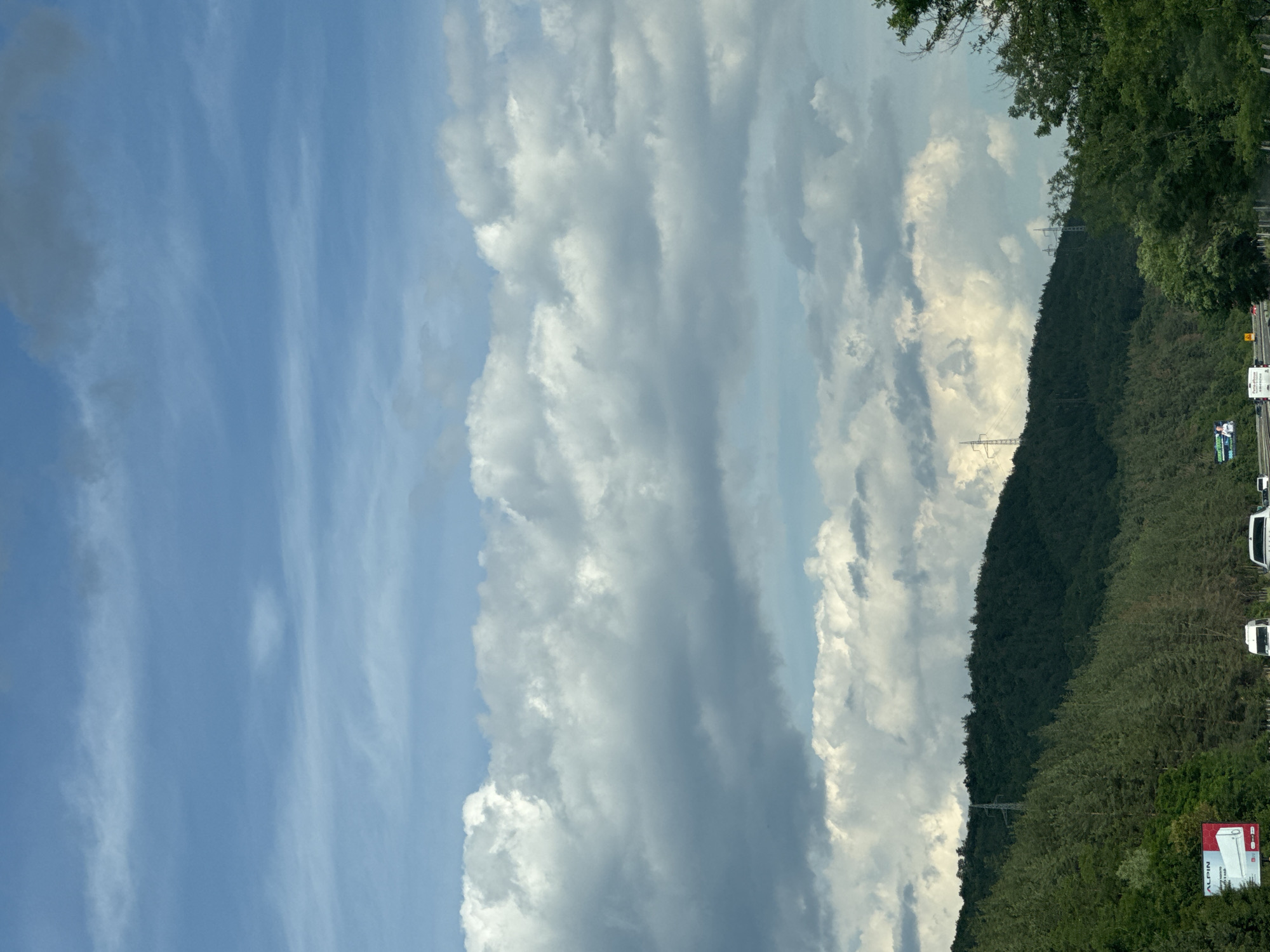 The image captures a scenic view of a mountainous landscape under a dramatic sky, with a highway passing through. The foreground features a road with several vehicles, including white vans and cars, indicating traffic. To the left, a billboard advertising "ALPIN" is visible, partially obscured by trees.
The terrain rises steeply on the left, covered in dense green coniferous forest. A power line tower is situated on the crest of the hill, with more such towers visible further in the distance. The sky is a prominent feature, filled with layers of cumulus clouds, ranging from bright white to dark gray, creating a dynamic and somewhat overcast atmosphere. The upper portion of the sky is a lighter blue with wispy cirrus clouds. The overall impression is a natural landscape with human infrastructure intersecting it, likely on the outskirts of Rajana, Bulgaria, given the context.