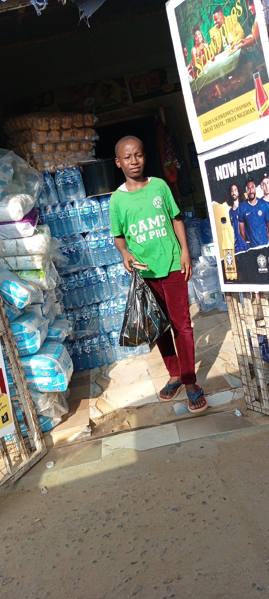 A young boy stands in front of a market stall in Sabon Gari East, Nigeria, holding a black plastic bag. He is wearing a bright green t-shirt with the words "CAMP IN PRO" printed on it in white and burgundy corduroy pants, along with blue and tan flip-flops. The stall is filled with stacks of bottled water and what appear to be packets of baby diapers. To the right of the boy, a large advertisement poster is displayed, featuring a group of men in blue soccer jerseys and a bottle of energy drink. The ground is tiled with a pattern of light and dark squares, and the overall lighting suggests an outdoor, sunny environment.