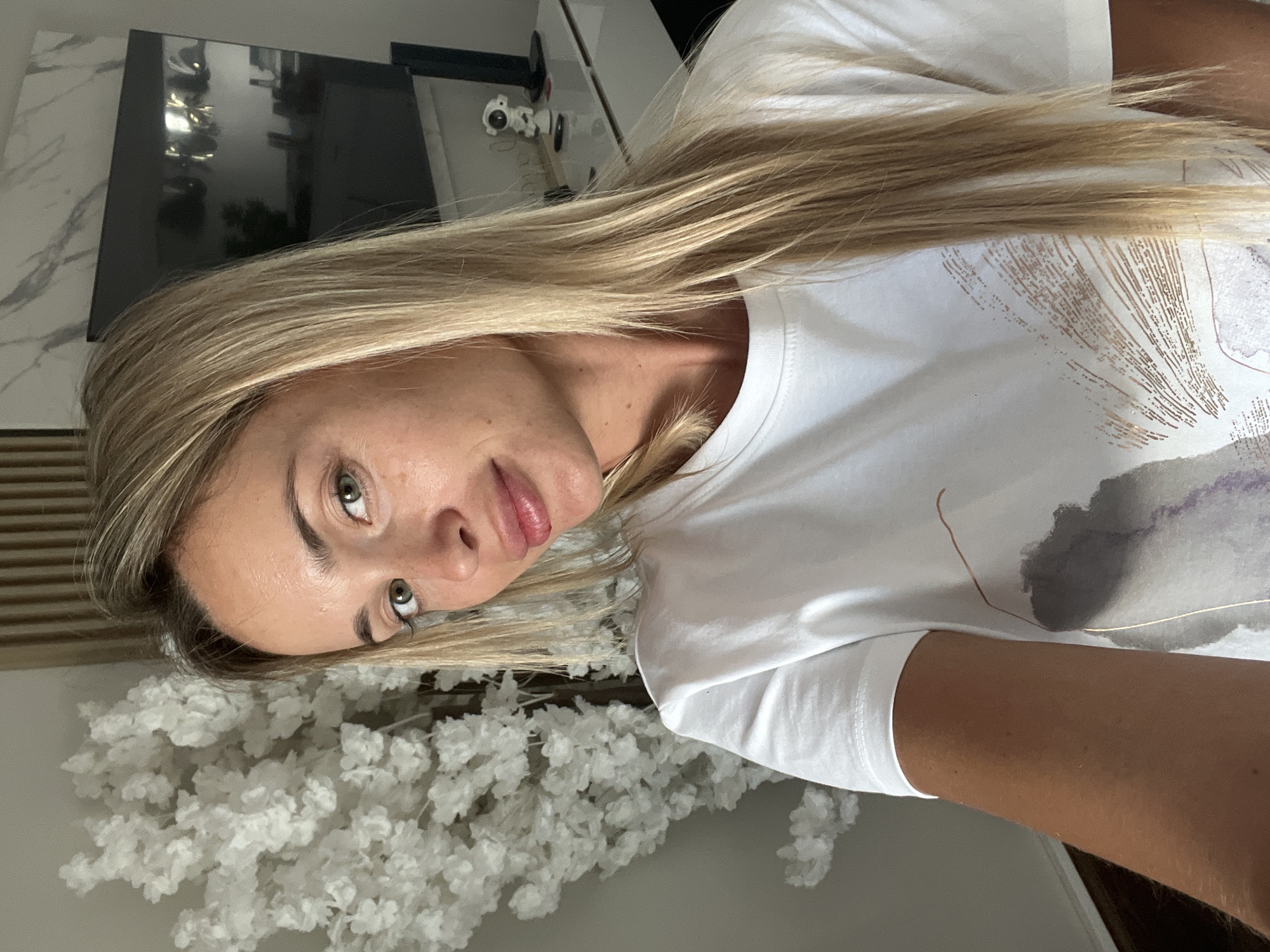 A blonde woman with light skin and green eyes is taking a selfie indoors. She is wearing a white t-shirt with an abstract design. Her hair is long and blonde. To her left is a large, artificial white floral tree. In the background, a large television is mounted on the wall, and a white dresser with some objects on it is visible. The overall setting appears to be a domestic interior, potentially a living room or bedroom. There are no indications suggesting this location is in Sharm El Sheikh, Egypt.