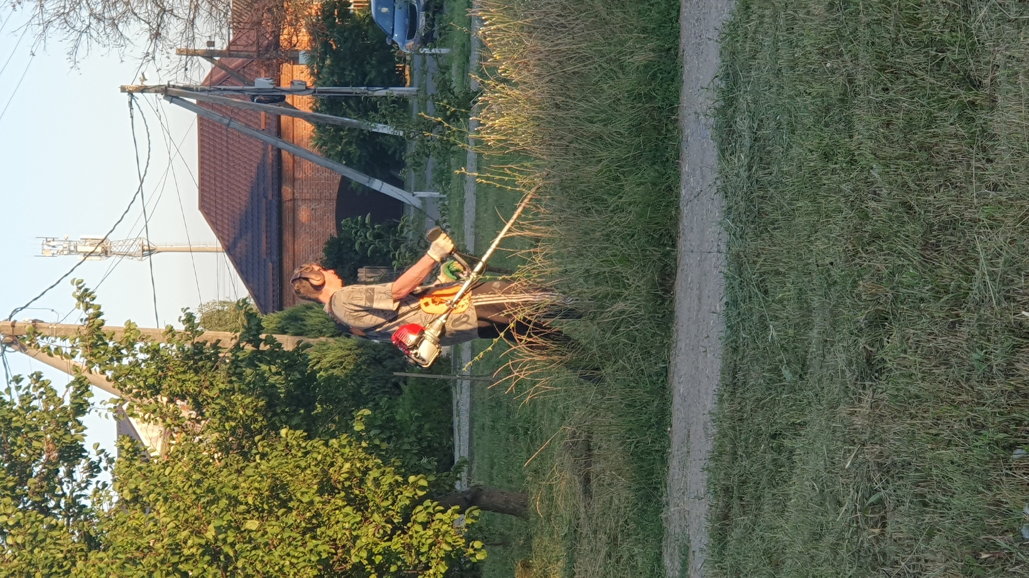In Ust-Labinsk, Russia, a person is seen trimming a grassy area with a string trimmer. The individual, wearing a patterned shirt, dark pants with a white stripe, gloves, and headphones, is positioned on a slightly elevated patch of land adjacent to a paved surface. Behind them, a tree with lush green foliage is visible, as are several buildings with brown roofs and brickwork. A tall cell tower stands in the distance, alongside utility poles and overhead wires. The lighting suggests it is either early morning or late afternoon, with the sun casting long shadows and highlighting the dry grass. A dark-colored car is parked on the right side of the frame.