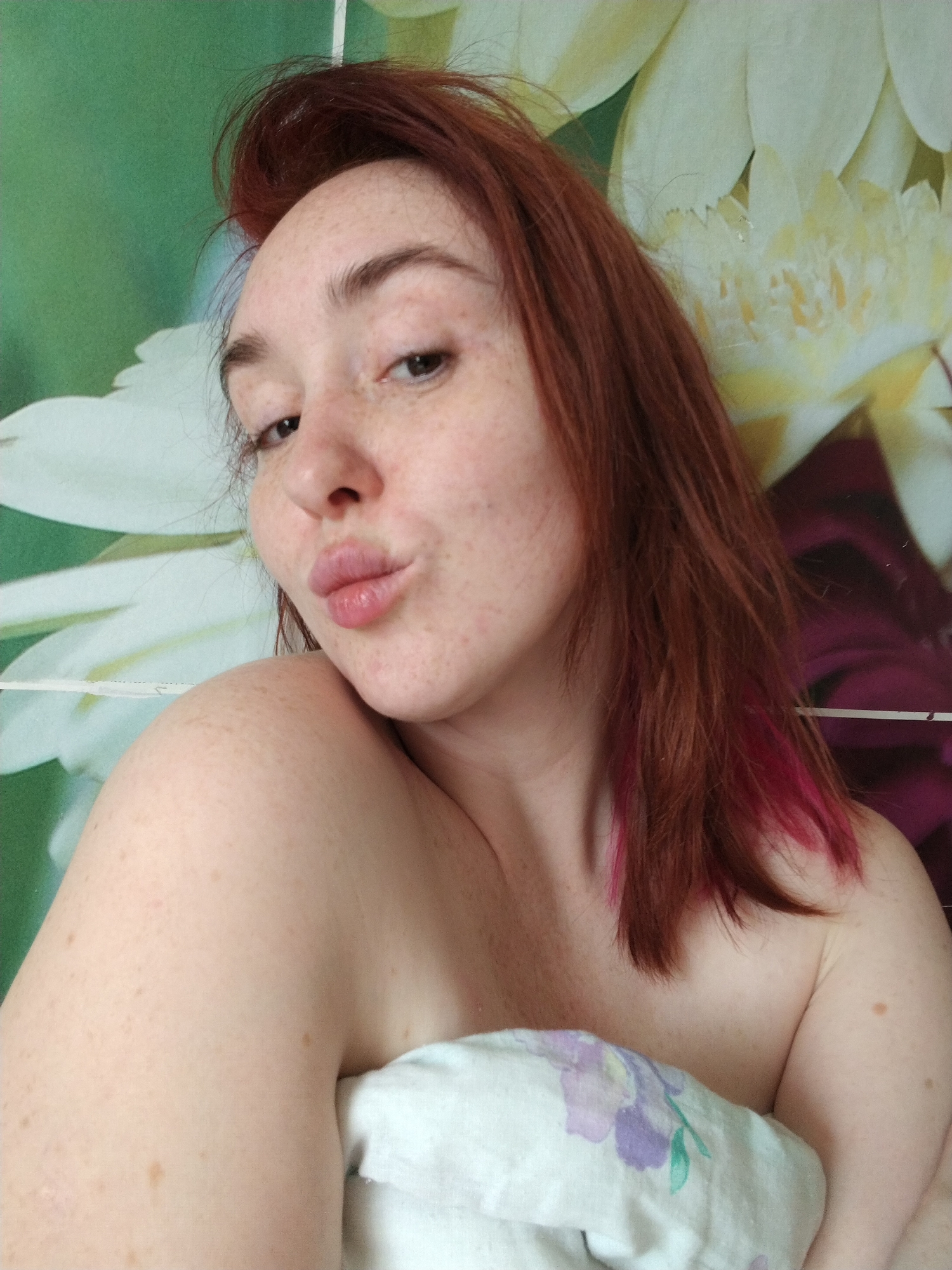 A person with fair skin, freckles, and reddish-brown hair with pink-tinted ends is posing for a close-up selfie. The person's hair is slightly disheveled, with some strands appearing to be sticking up. The person is making a "kissing" facial expression, with their lips pursed. The person is partially covered by a white sheet with a floral pattern. The background consists of a light green wall and a design of white flowers. No evidence suggests this photo was taken in Krasnogorskoye, Russia.