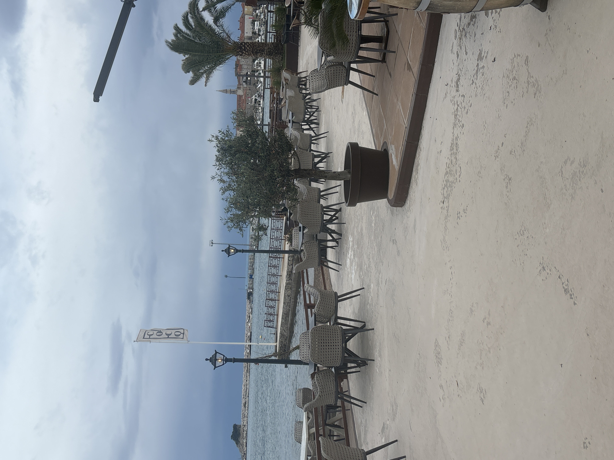 The image depicts an outdoor dining area in Budva, Montenegro. Rows of empty tables and chairs are arranged along a waterfront promenade. The tables are set with wicker chairs, and the area is decorated with trees in large pots. In the background, the sea is visible, with a small island in the distance. The sky is overcast. In the further background is a view of Budva's old town.