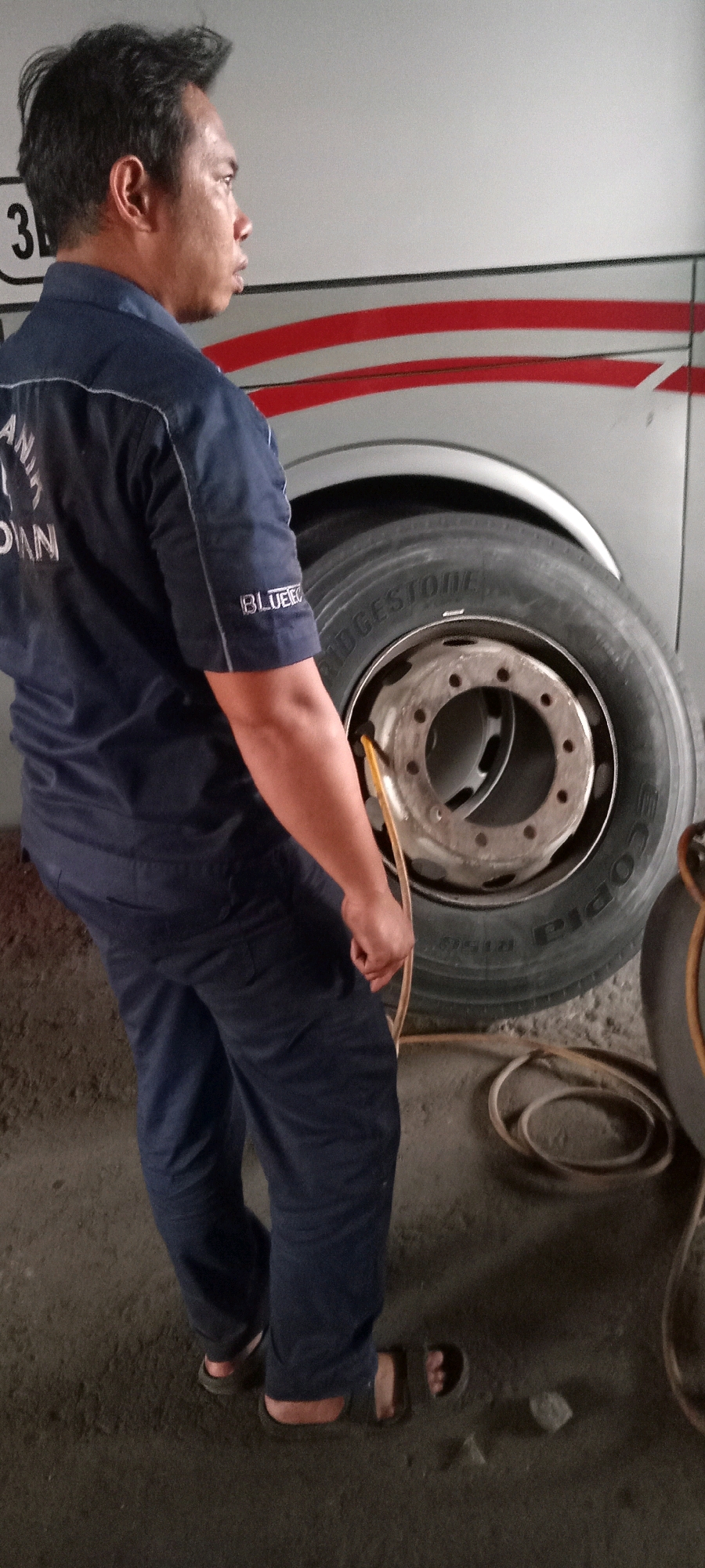 A side-view medium shot captures a man in a dark blue mechanic's uniform, standing next to a large truck tire. He is positioned to the left of the frame, facing the right. His uniform has white lettering on the left chest and sleeve, with the word "BLUEE" visible on the sleeve. The man is holding a yellow air hose connected to the tire valve. The tire itself is a Bridgestone brand, with prominent tread. The truck it's attached to has a silver exterior with two red horizontal stripes. The ground appears to be a rough, dirt-like surface. In the background, another tire is partially visible to the right of the main subject. The scene suggests a mechanic at work, likely in a garage or service area. The number "3" is visible on a white background near the top left of the frame.