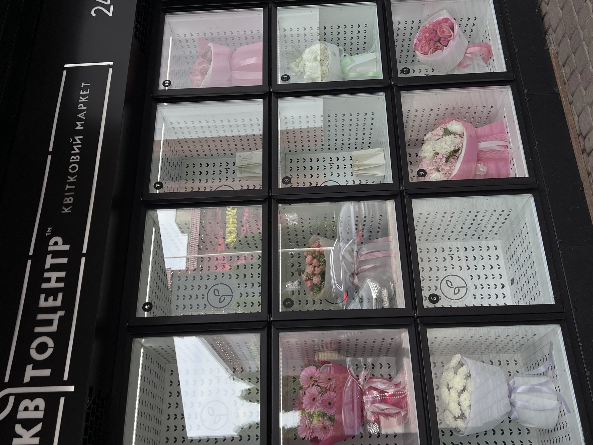 The image displays a modern, black outdoor flower vending machine or self-service flower locker system. It features a grid of twelve transparent, square compartments, arranged in three columns and four rows.
Inside several compartments, various pre-arranged flower bouquets are visible. These include pink roses in pink wrapping (compartments 10 and 12), white flowers in white and light green wrapping (compartment 11), a mixed bouquet of pink and white flowers (compartment 9, second row from top), pink gerberas accompanied by a small red toy car (compartment 5), and white chrysanthemums or daisies in white and lavender wrapping (compartment 9, bottom row). Some compartments are empty, revealing a white interior patterned with small leaf shapes and a stylized plant logo. Each occupied compartment is labeled with a circular number.
The machine's left side features text in Ukrainian: "КВІТОЦЕНТР" (Flower Center), "КВІТКОВИЙ МАРКЕТ" (Flower Market), and the number "24," likely indicating 24-hour service. The setting appears to be an urban outdoor area during daytime, with paving stones visible on the ground to the right. The image suggests an automated, convenient way to purchase fresh flowers in Ternopil, Ukraine.