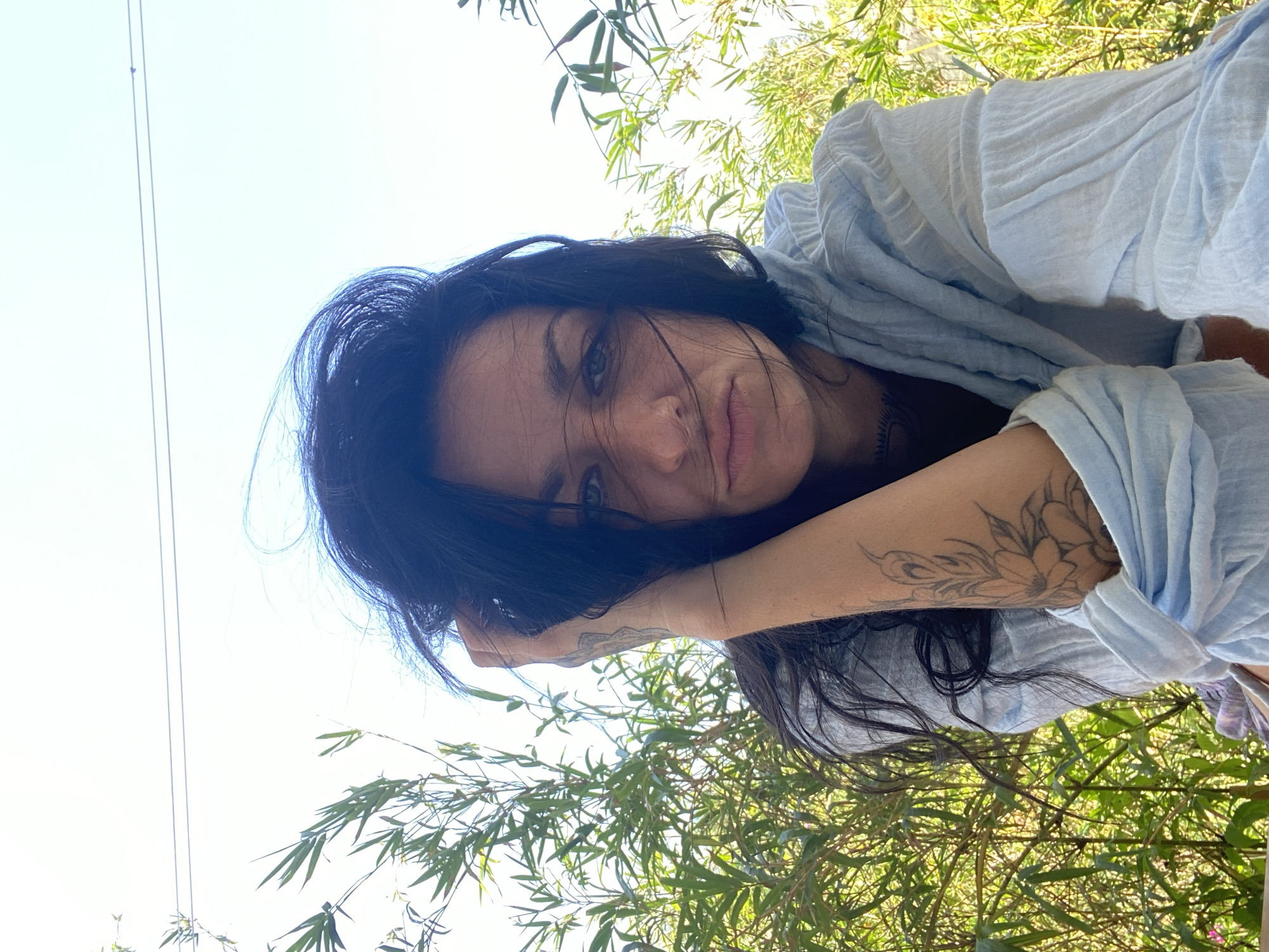 The image shows a woman with dark hair and light eyes lying down outdoors. She is wearing a light blue, loose-fitting top. Her left arm is visible, featuring a floral tattoo. The background consists of green foliage, possibly bamboo, and a bright blue sky. The location is potentially Mũi Né, Vietnam, given the prompt's instruction.