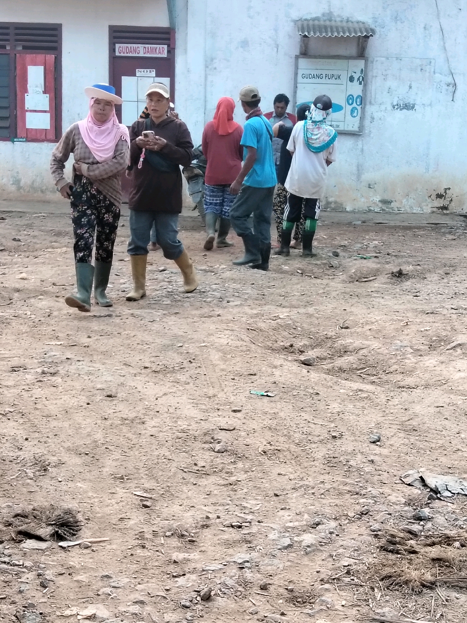 In the city of Teluk Tenggirik, Indonesia, a group of individuals are gathered outdoors in front of a building. The building has white walls and two doors visible. Above one door, signage reads "GUDANG DAMKAR" and "SOP". Above another doorway on the right side of the building, a sign reads "GUDANG PUPUK" and displays several symbols. The ground in front of the building is unpaved and appears to be dirt and gravel, with some debris scattered around.
Two women in the foreground are walking from left to right. The woman on the left wears a pink headscarf, a patterned long-sleeved shirt, and dark patterned pants. She is also wearing tall green boots. The woman next to her wears a beige cap, a brown hooded jacket, and blue jeans. She is holding a mobile phone in her right hand and is wearing tall beige boots.
Behind them, several other individuals are visible. One person wears a red headscarf and a red long-sleeved shirt, with patterned shorts. Another person in a blue t-shirt and dark pants walks away from the camera. Other figures are partially obscured. The environment appears to be utilitarian, possibly associated with storage or agricultural facilities.