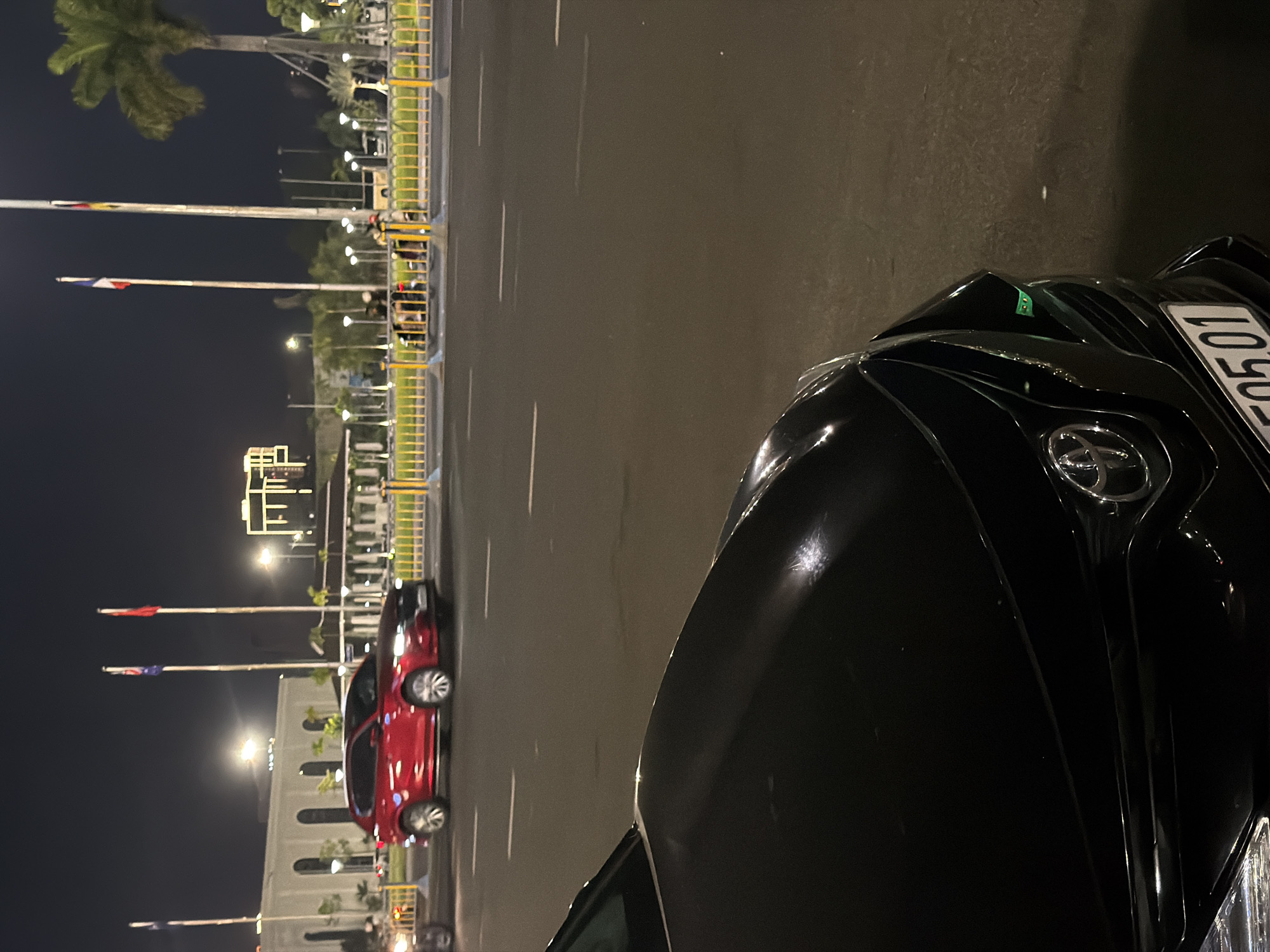In Đà Nẵng, Vietnam, at night, a black Toyota car is parked on a city street. The foreground is dominated by the hood and grille of the black car, revealing its Toyota emblem and part of its license plate, "0501." In the middle ground, a red SUV is driving on the same road, with a yellow barrier and some palm trees visible behind it. The background shows buildings illuminated with lights, along with flags on poles.