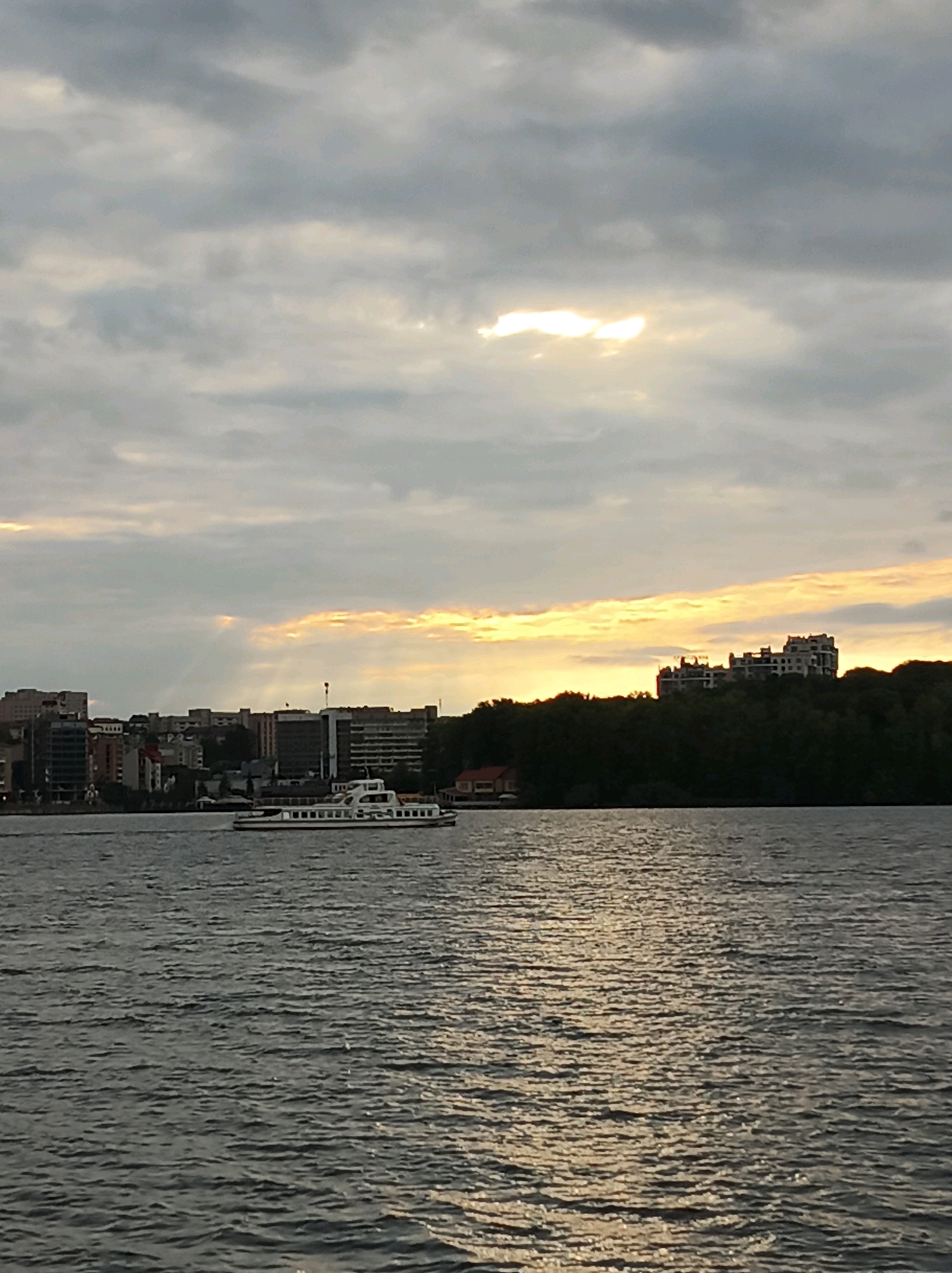 The image depicts a wide body of water with ripples, likely a lake or river, with a city skyline in the background under a cloudy sky. A white passenger boat with multiple windows and a dark roof is sailing on the water, positioned in the middle ground, slightly to the left of the frame. The city skyline features a collection of buildings of varying heights and architectural styles, with some appearing to be apartment buildings or office complexes. Trees form a dark band of foliage along the top of the city skyline, particularly on the right side, where a large, multi-story building is situated on a hill overlooking the water. The sky is filled with dramatic clouds, with patches of sunlight breaking through in the upper part of the image, creating a diffused, atmospheric lighting effect, and streaks of sunlight are also visible filtering through the clouds towards the horizon. The overall scene suggests a serene, late afternoon or early evening ambiance in an urban environment situated near a natural waterway. The location is Ternopil, Ukraine.