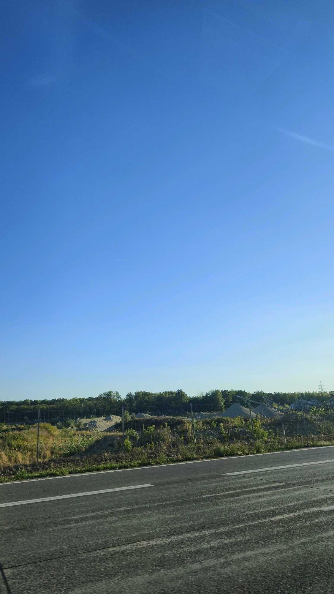The image depicts a bright, clear blue sky with a faint contrail visible in the upper portion. Below the sky, a landscape features a line of trees forming a dense, green horizon. In front of the trees, a fence runs horizontally, separating the foliage from a sandy, disturbed ground area with several mounds. A paved road with a white lane marker occupies the foreground, suggesting a travel or roadside perspective. The scene appears to be an outdoor setting, possibly along a highway or a rural road, with a construction or industrial site suggested by the mounds of material and the fence. The overall impression is one of daytime, with good visibility and a vast expanse of sky. The location is identified as Spillern, Austria.