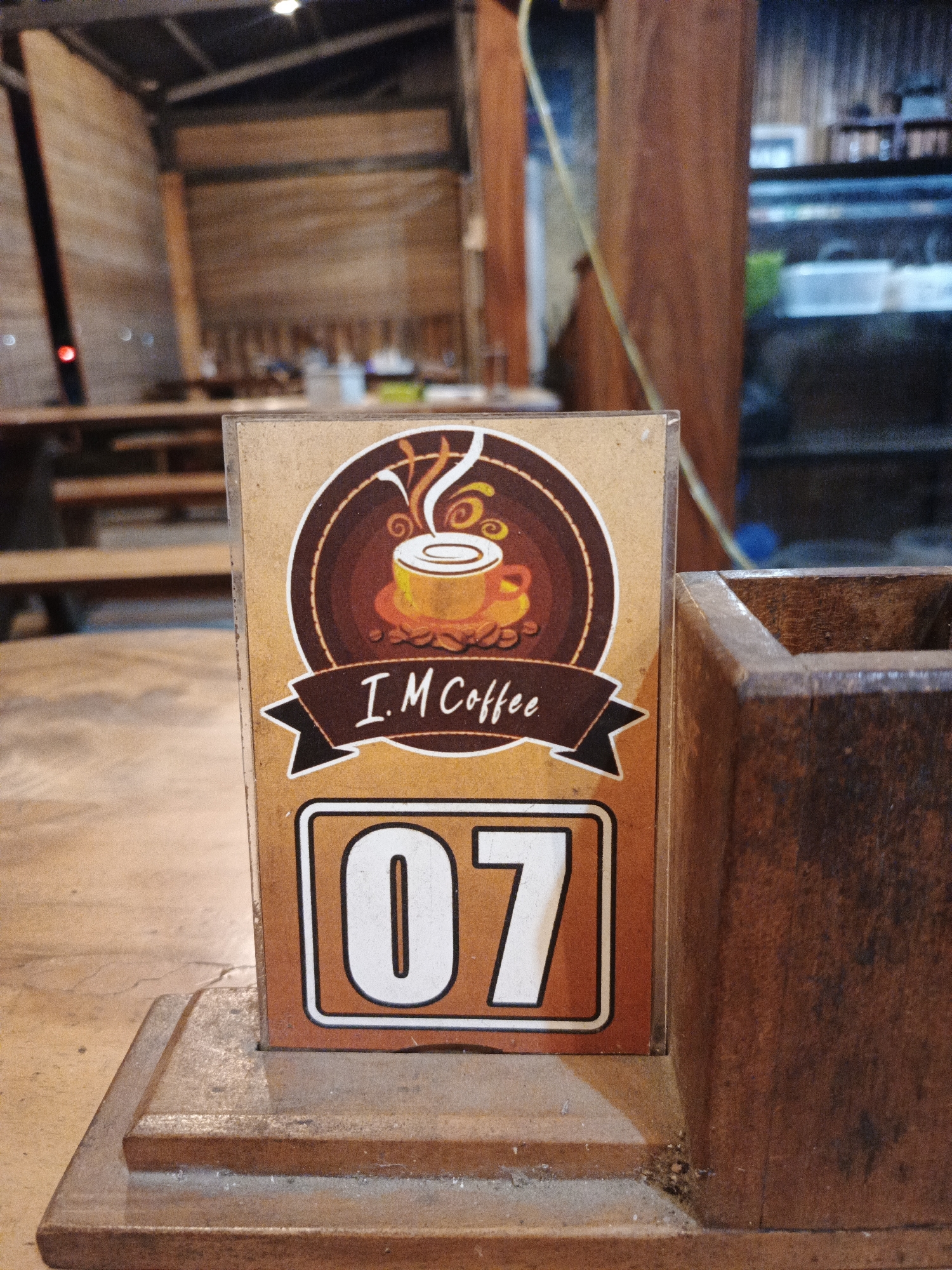 A close-up shot focuses on a table number sign, marked '07', for 'I. M Coffee' in Geulumpangmeujim-Jim, Indonesia. The sign features a brown and yellow coffee cup logo. It is placed on a worn wooden surface next to a wooden container, likely a utensil or condiment holder. In the blurred background, one can discern a wooden structure, potentially a part of the cafe's interior or a surrounding building. Seating is visible in the background.