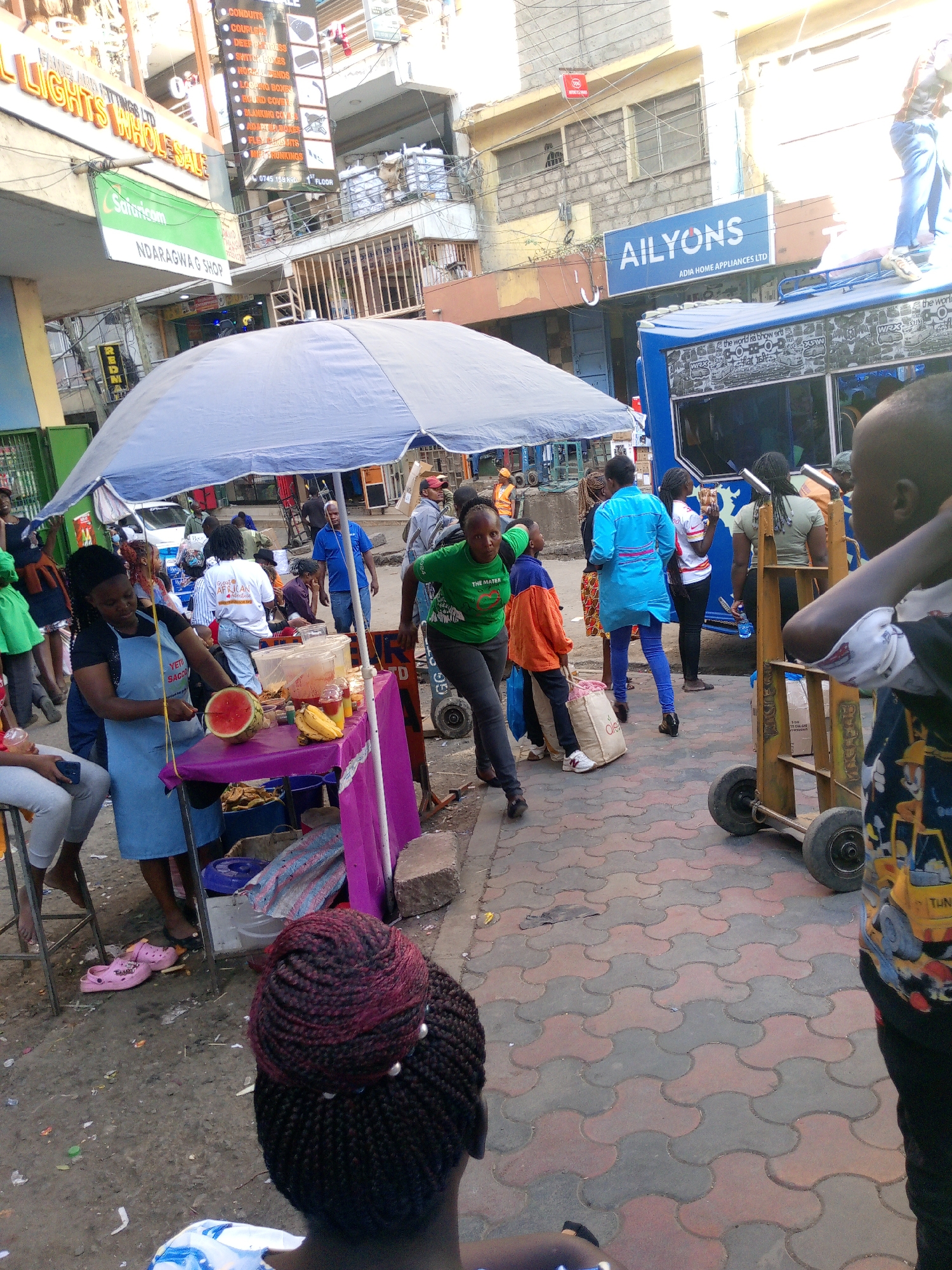 The image captures a vibrant street scene in Nairobi, Kenya. A makeshift fruit stand, shaded by a large blue umbrella, is situated on the sidewalk, with a woman in a blue apron preparing a watermelon. Various individuals surround the stand, some appearing to be customers, while others walk past.
In the background, buildings with signs like "AILYONS ADIA HOME APPLIANCES LTD," "ALL LIGHTS WHOLESALE," and "Safaricom NDARAGWAG SHOP" can be identified. A blue vehicle is parked nearby, and a hand truck loaded with packages is present. The street is paved with interlocking grey and red blocks. A person wearing a shirt with a cartoon truck on it is in the foreground, partially obscuring the view.
