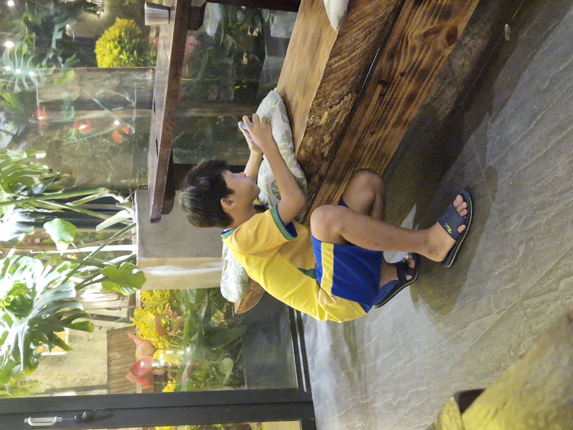 A young boy in a yellow and blue shirt and shorts is playing a video game on a mobile phone. He is crouching on the floor next to a rustic wooden bench in what appears to be a cafe or restaurant. The boy is holding a white pillow with green and white patterns for support. The background shows a large window with lush green plants, some of which appear to be Monstera leaves, and a wooden table and chairs. The flooring is a dark gray, textured material. The lighting suggests it is indoors, possibly at night. The overall scene is one of leisure and entertainment. This cafe is located in Da Nang, Vietnam.
