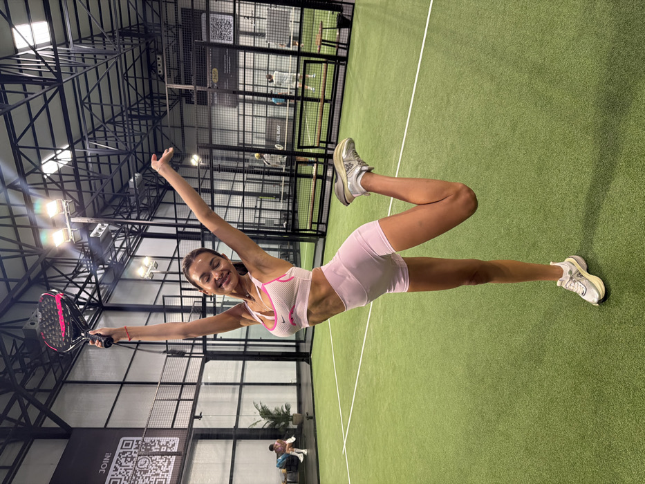 The image displays an indoor padel court facility located in Choeng Thale, Thailand. A female subject with long brown hair stands centrally on a green artificial turf court marked with white lines. She wears a pink sports bra with white accents, light pink high-waisted shorts, and silver-white athletic shoes. Her pose involves standing on her left leg, with her right leg bent and foot raised behind her. Both arms are extended overhead, her left hand holding a black and pink padel racket. Her head is tilted, and her eyes are closed, indicating a smile. The court perimeter consists of transparent glass panels and black metal fencing. Overhead, multiple industrial light fixtures illuminate the area, mounted on exposed black metal trusses of the ceiling structure. A black banner featuring a 'JOIN!' QR code is visible on the far left wall. In the background, other individuals are present on adjacent padel courts, separated by fencing and nets, with one person seated on a bench on the left side of the frame near a potted plant.