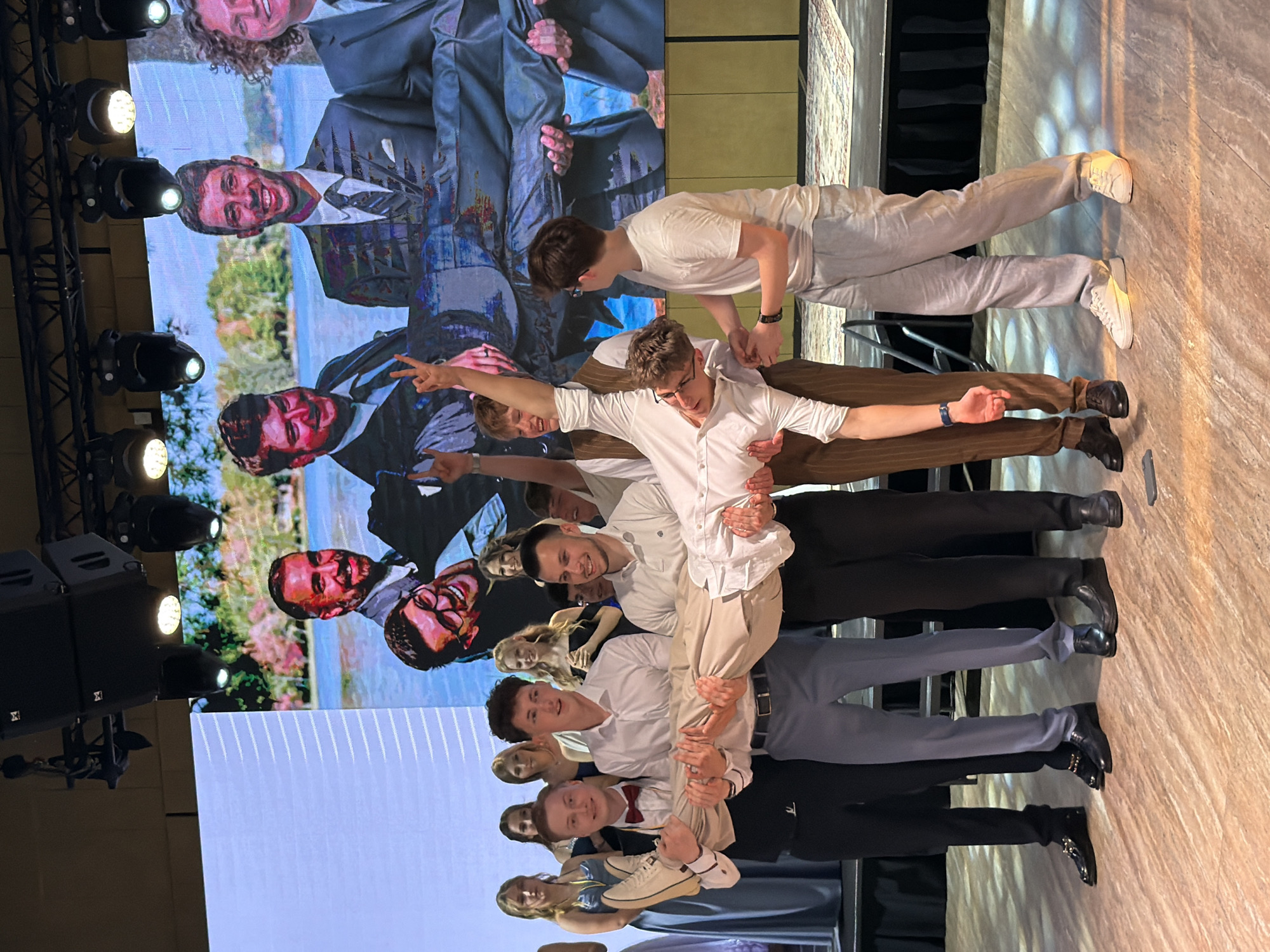 This is a medium shot photograph of a group of young people, predominantly males in formal attire, on a stage in front of a large screen displaying an image of people in formal wear by a body of water. In the foreground, four young men are carrying another young man horizontally, who is wearing glasses and a white shirt. The young man being carried is looking towards the viewer, with his head tilted back and arms outstretched. The men carrying him are dressed in dress shirts and slacks of varying colors. Behind this group, several young women in formal dresses and a few young men in suits are standing, some with their hands raised. The stage appears to be set up for an event, possibly a prom or formal gathering. The backdrop is a large LED screen showing a digitally rendered or photographed image. Lighting is directed at the subjects on stage. The floor is a polished wooden surface. The overall scene suggests a celebratory or performance-related event. There is no indication that this image is located in the city of Khotianivka, Ukraine.