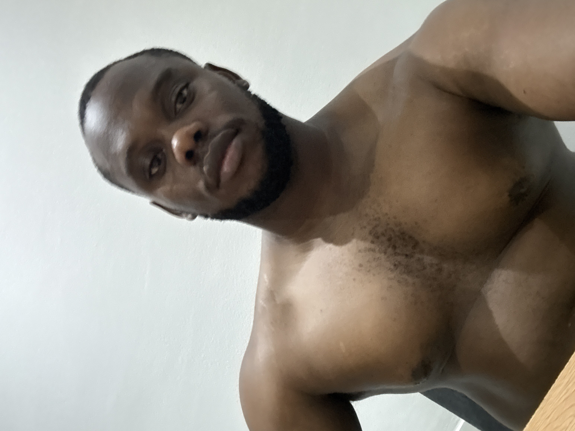 An indoor, low-angle portrait captures a muscular Black man looking directly at the camera. He has short dark hair and a neatly trimmed beard. The man is shirtless, showcasing his well-defined chest and shoulders, with some dark chest hair visible. His head is tilted slightly, and he maintains a neutral, direct gaze. There are distinct lighter skin patches visible on his chest and upper arm. The background is a plain, light-colored wall, and a hint of a light wooden surface is visible in the bottom right corner of the frame. The lighting appears soft and even, suggesting an indoor setting without natural sunlight. There is no visible text in the image. The setting is Iddo Tudunwada, Nigeria.