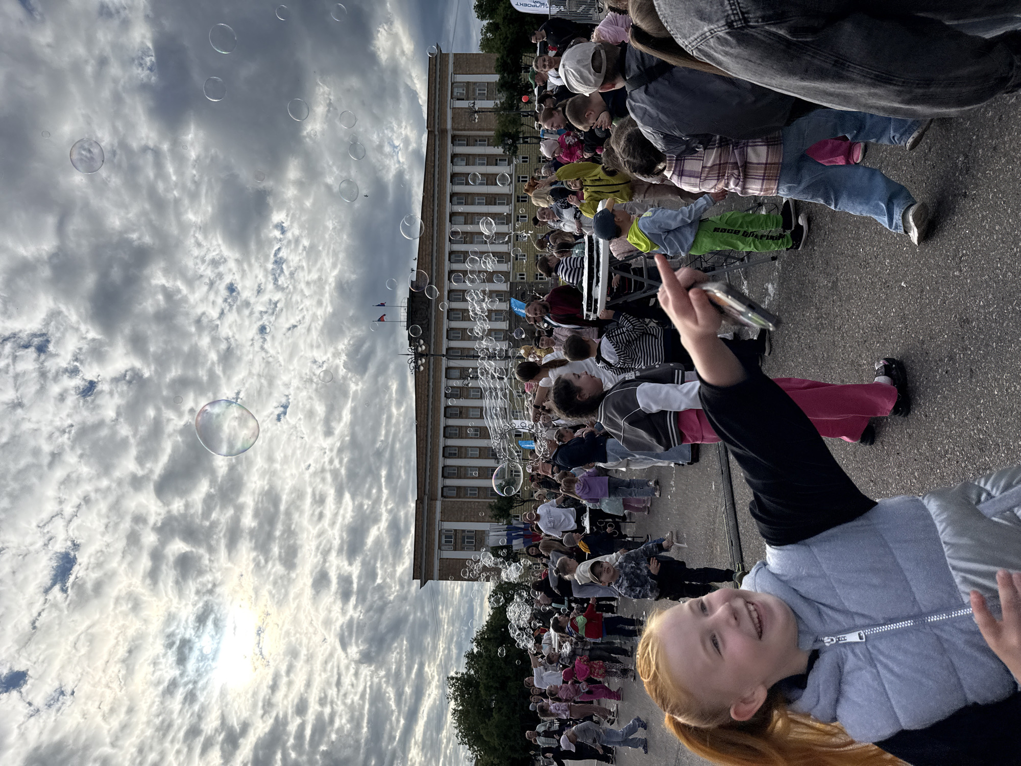 A slightly angled full outdoor shot shows a crowd of children and adults in front of a large building in what appears to be a city square. Numerous bubbles float in the air. In the foreground, a red-haired girl is seen smiling and looking toward the crowd, possibly taking a photo or video with her phone. She wears a black long-sleeved shirt and a light-gray puffer vest.
Behind the girl, a large group of people, mostly children, are standing or moving slightly. Some are looking up at the bubbles. Behind the crowd is a large, multi-story building with many windows. The sky is overcast with many cloud formations. In the left corner of the sky, the sun is shining slightly through the clouds. A few trees are also visible behind the building.
The location is likely the square in front of the administration building on Ploshchad Pobedy-Sofiyskaya in Veliky Novgorod, Russia, based on the architecture of the building in the background.