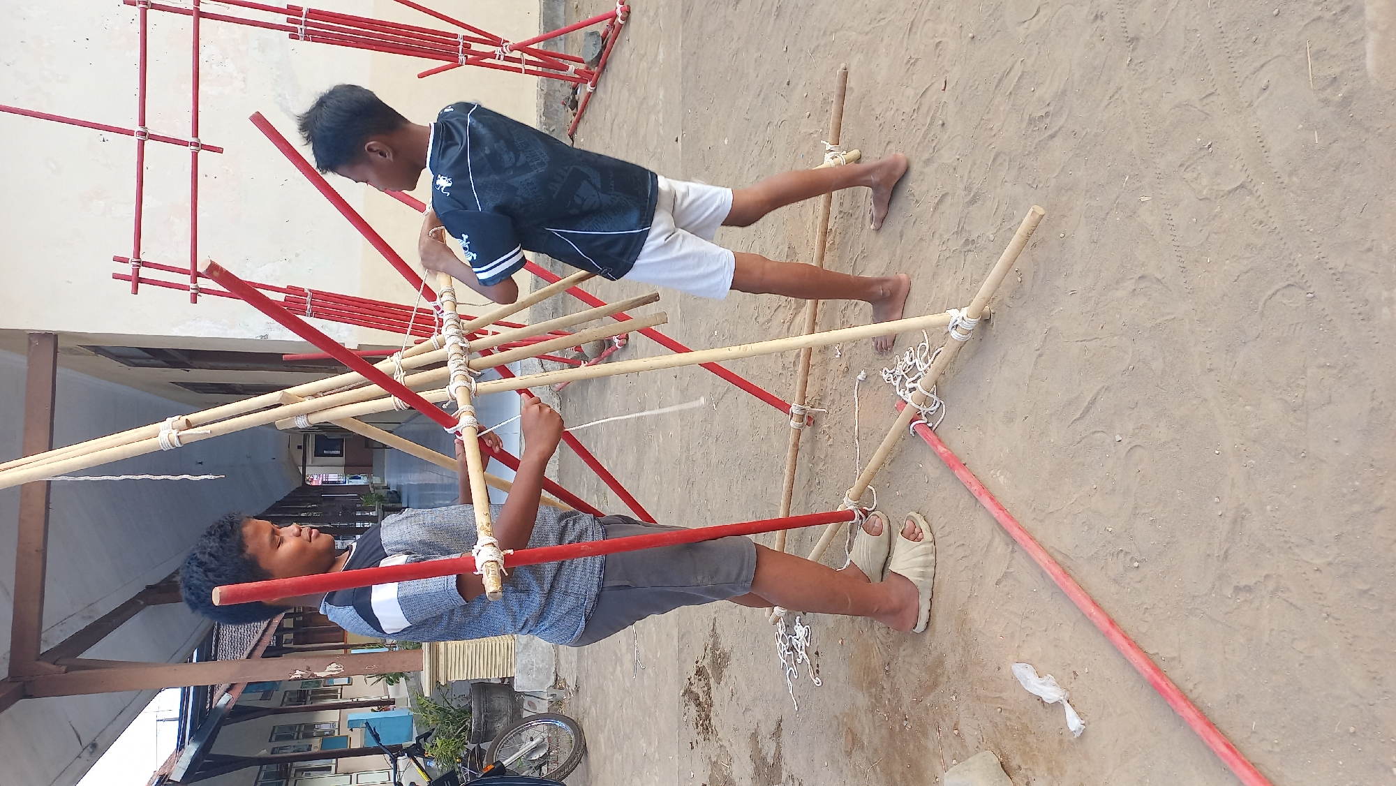 Two young boys are engaged in an outdoor activity involving bamboo poles and red painted poles. They are standing on sandy ground in front of a building with a covered walkway. One boy, on the left, wears a gray and white striped t-shirt and grey shorts, with beige sandals on his feet. The other boy, on the right, is in a dark blue t-shirt with white detailing and light grey shorts, barefoot. They appear to be constructing or manipulating a structure made of these poles, tied together with white rope. In the background, the building has a light-colored facade and windows. There is also a motorcycle visible to the left, partially obscured. The scene suggests a schoolyard or an outdoor community area in Mangunsari, Indonesia, possibly during a scout or outdoor education activity.