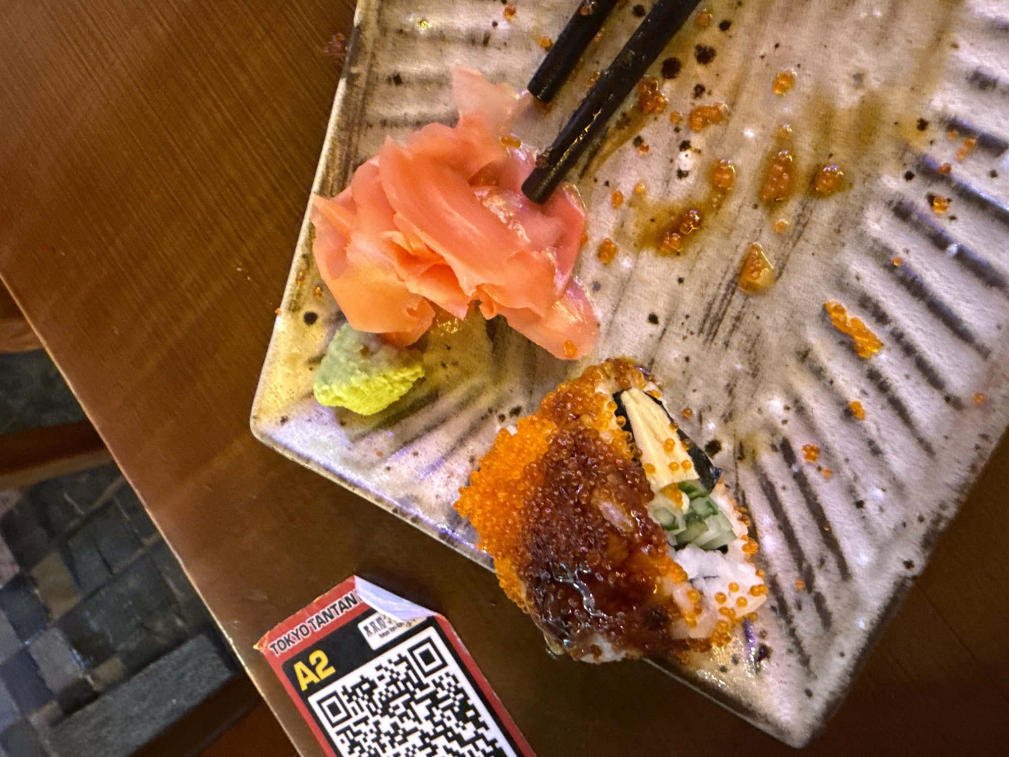 The image depicts a nearly finished plate of sushi at what appears to be a restaurant in Hà Nội, Vietnam. A portion of a wooden table is visible, along with part of a chair with a patterned seat. The plate, likely made of ceramic, is mostly light gray with dark gray stripes and scattered food remnants. A small pile of pink pickled ginger and a dollop of green wasabi are on the upper left. A pair of black chopsticks rests partially on the plate. A single piece of sushi, covered in orange roe and a dark brown sauce, is positioned near the bottom center of the plate. A small, red and white ticket labeled "TOKYO TANTAN A2" with a QR code is placed near the sushi. The plate also contains scattered droplets of orange sauce and dark brown spots. The scene suggests a recently consumed meal in a restaurant setting.