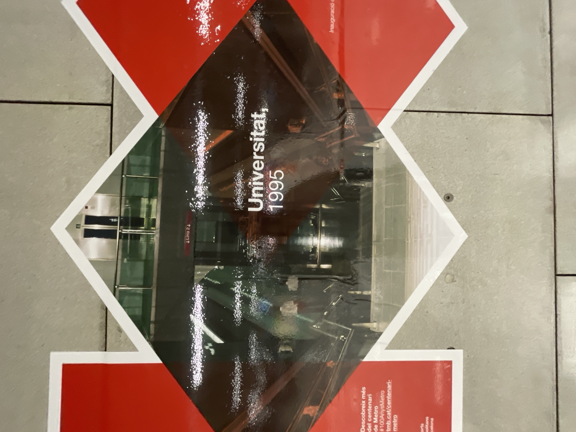 The image shows a promotional floor sign, likely at a Barcelona Metro station. The sign is diamond-shaped with a red border and white inner lines. Inside the diamond is a black and white image of the Universitat Metro station interior, including train tracks and a platform area with people waiting for the train. The text "Universitat, 1995" is displayed across the image, suggesting the station's inauguration date or a significant event. Smaller text below the image promotes the Metro's centennial, and the text, "#100AnysMetro tmb.cat/centenari-metro" is also present on the poster. "Linia 2" is on the upper left of the image. The sign is placed on a tiled floor.