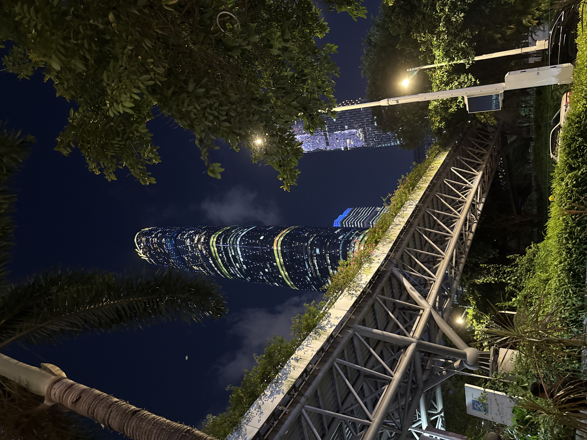 This photograph, taken at night, shows a view looking upwards in the Tianhe District, China. Visible in the scene are two skyscrapers illuminated with lights. One is taller and more cylindrical in shape than the other. A large elevated pedestrian walkway is present, with plants growing on its sides. The steel structure of the walkway is visible. Streetlights shine nearby. A palm tree is in the foreground, obscuring part of the taller skyscraper. Branches from other trees are visible at the top of the frame.