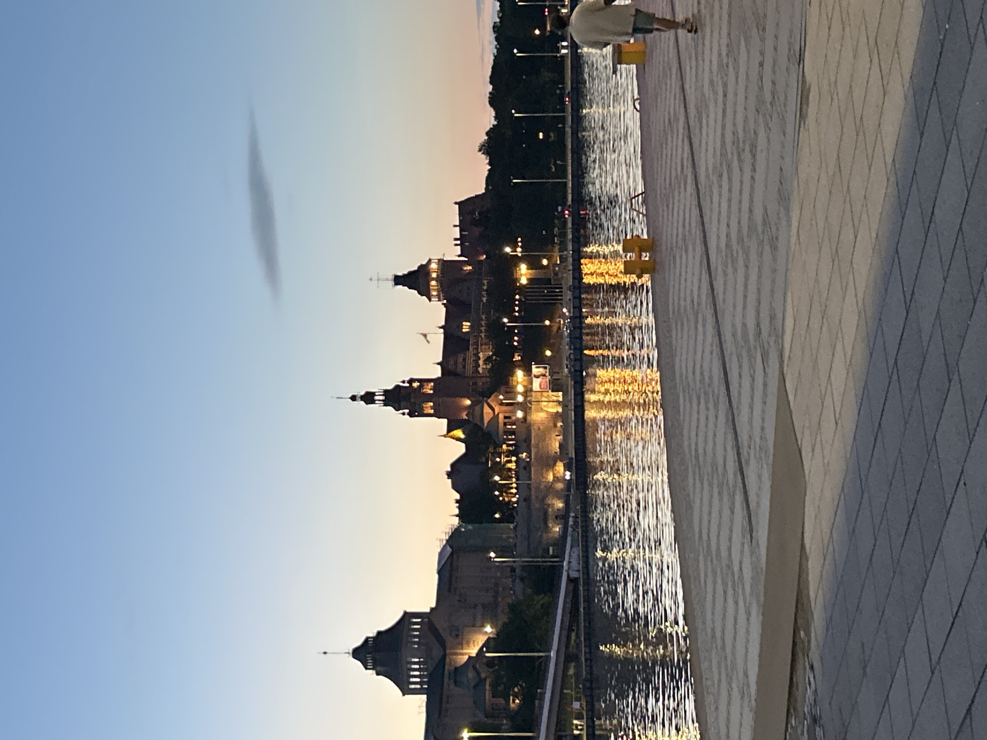 A wide-angle, eye-level shot captures a waterfront scene in Szczecin, Poland, at dusk. The sky is a gradient of pale blue to pink. In the background, a grand building complex with multiple spires and illuminated windows is visible across a body of water. The building reflects warmly on the rippling water's surface. A bridge extends from the left side of the frame towards the building. The foreground is dominated by a paved walkway made of interlocking grey and tan square tiles. A single individual, a person wearing a light-colored shirt and shorts, walks away from the camera on the walkway towards the right. Yellow bollards are positioned along the edge of the walkway. Streetlights are visible along the waterfront.