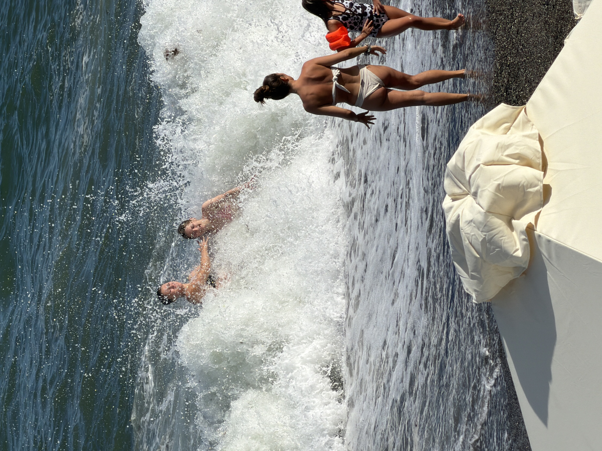 The image depicts a dynamic scene at a beach in Alushta, Ukraine, with a large wave crashing. In the water, two individuals are partially submerged and caught within the crest of the wave, appearing to be enjoying the splash. To the right, on the shore, two females are standing. One, in a beige bikini, faces away from the camera with her back to the ocean. The other, a child wearing a leopard-print swimsuit and orange arm floaties, stands beside her, also facing away. In the foreground, a portion of a large, cream-colored beach umbrella is visible, casting a shadow on the dark, pebbly shore. The sea is a deep blue-green, with white foam and spray from the breaking wave dominating the mid-ground.