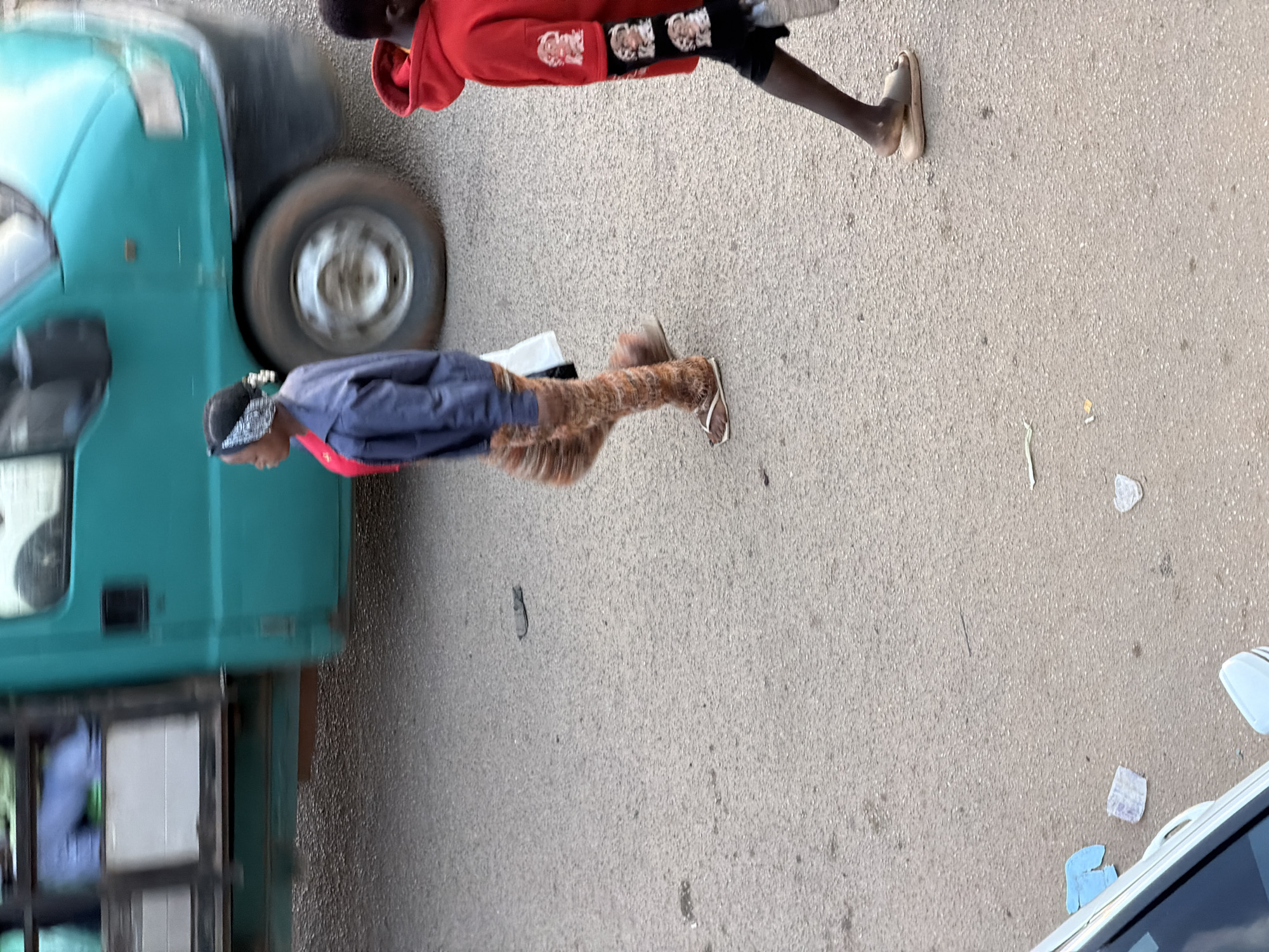 In Bukuru, Nigeria, two individuals are walking on a paved road beside a teal truck. On the left, a person wearing a dark blue jacket and patterned leggings walks with their body facing left and head turned towards the viewer. They are also wearing white flip-flops. To their right, a child in a red hooded sweatshirt and black pants with white graphic designs on the sleeves walks away from the viewer, also wearing a flip-flop on their right foot. The truck appears to be in motion, indicated by motion blur on its tires and body. The road surface is asphalt and there is some debris scattered on it, including pieces of paper.
