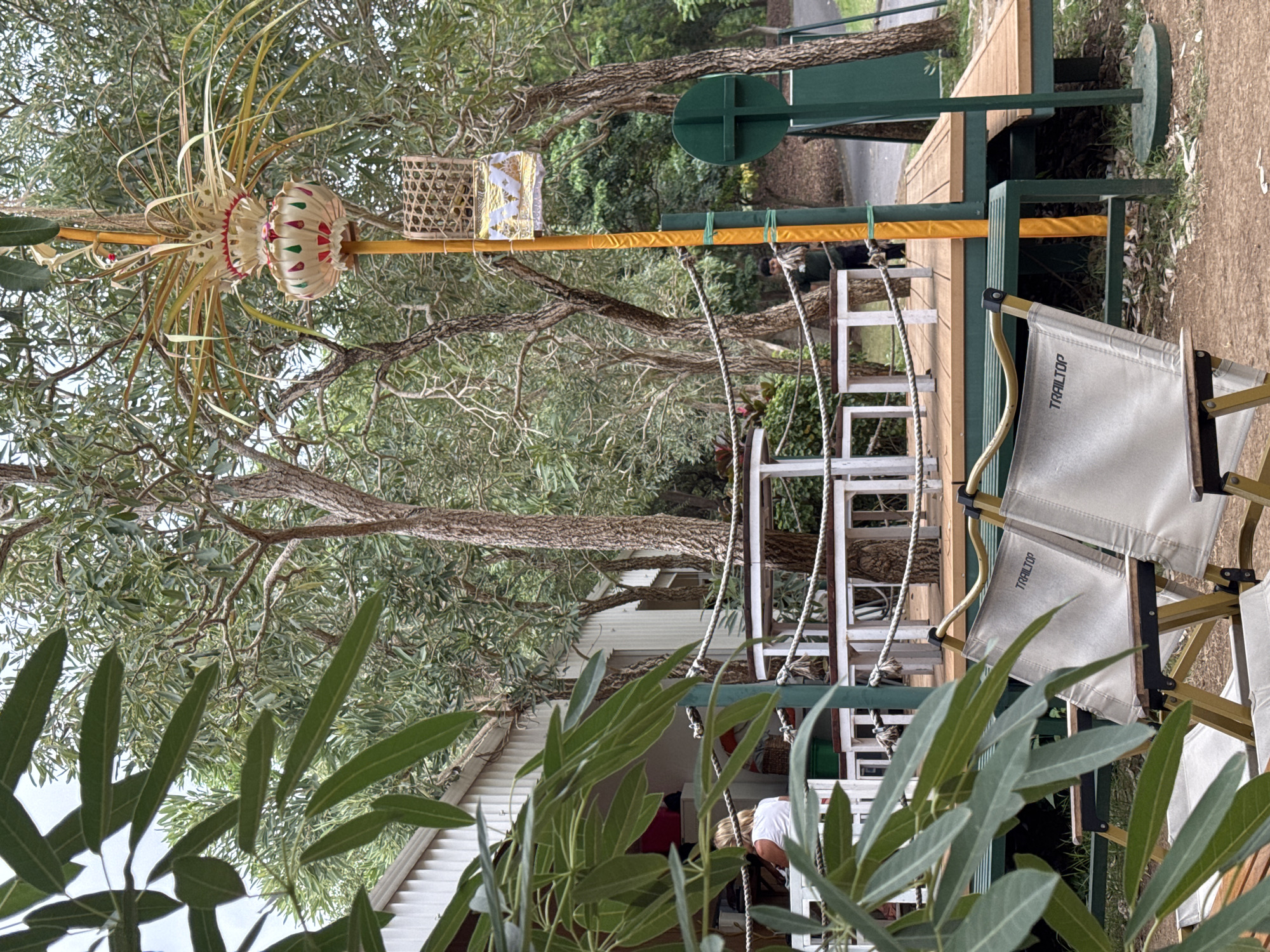 The image depicts an outdoor seating area with a thatched roof structure and a wooden deck. Several white tables and chairs are arranged on the deck, some with rope railings. Two portable camping chairs with the brand "TRAILTOP" are in the foreground. A yellow pole stands to the right, adorned with decorative items, including a woven basket and fabric elements. In the background, trees with textured bark and green foliage surround the area. A white building with a corrugated roof is partially visible on the left. A person in a dark shirt is standing near the back of the deck. The ground is covered in dirt and fallen leaves. The scene appears to be a casual outdoor dining or gathering space, possibly at a resort or cafe. The overall atmosphere is natural and relaxed. The location is inferred to be Kuta Selatan, Indonesia based on the architectural style and tropical vegetation.