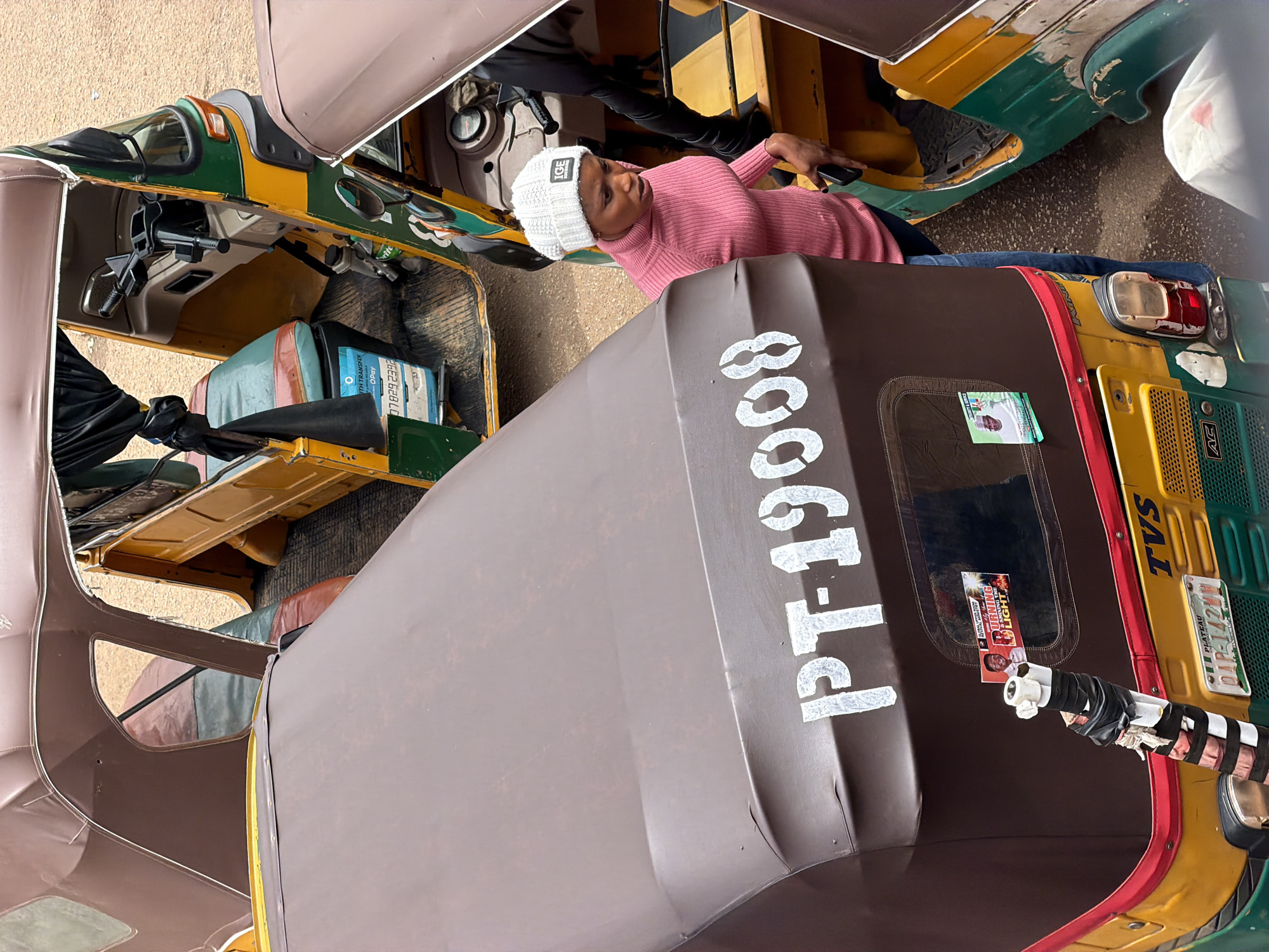 This media file shows a close-up, rotated view of three auto-rickshaws parked next to each other. The front auto-rickshaw is predominantly brown, with a dark brown canopy featuring the white stenciled number "PT-19008". A portion of the driver's side of this auto is visible, with a piece of tape-wrapped tubing attached to its side. To the left, another auto-rickshaw is visible, primarily colored green and yellow, with a brown and beige interior. A person wearing a pink sweater and a white knit cap is seated in this auto-rickshaw, holding a mobile phone and looking upwards. The third auto-rickshaw, partially visible on the right, is a bright green and yellow. The scene appears to be set on a gravelly or sandy surface, suggestive of an outdoor, possibly informal parking area. A small political sticker or flyer with an image of a person and text reading "BURNING LIGHT" and "SHINING THE" is affixed to the canopy of the brown auto-rickshaw. License plates on the green and yellow auto read "PLATEAU" and "011-1142". The lighting suggests it could be daytime. The overall impression is one of urban transportation in Nigeria, likely in a busy or roadside setting.