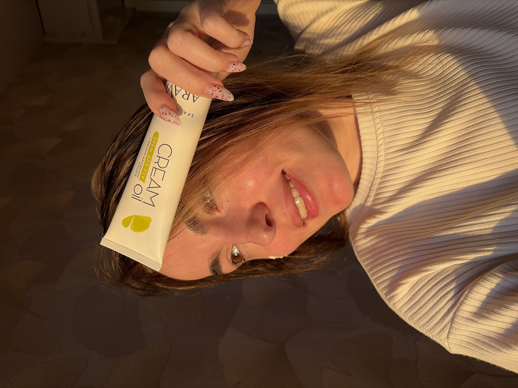 A woman is holding a tube of cream in front of her face. She is wearing a light-colored ribbed sweater. The tube is white with the words "CREAM OIL" written on it. The woman has brown hair and her nails are painted with a pink and red design. The image is taken in Izhevsk, Russia.