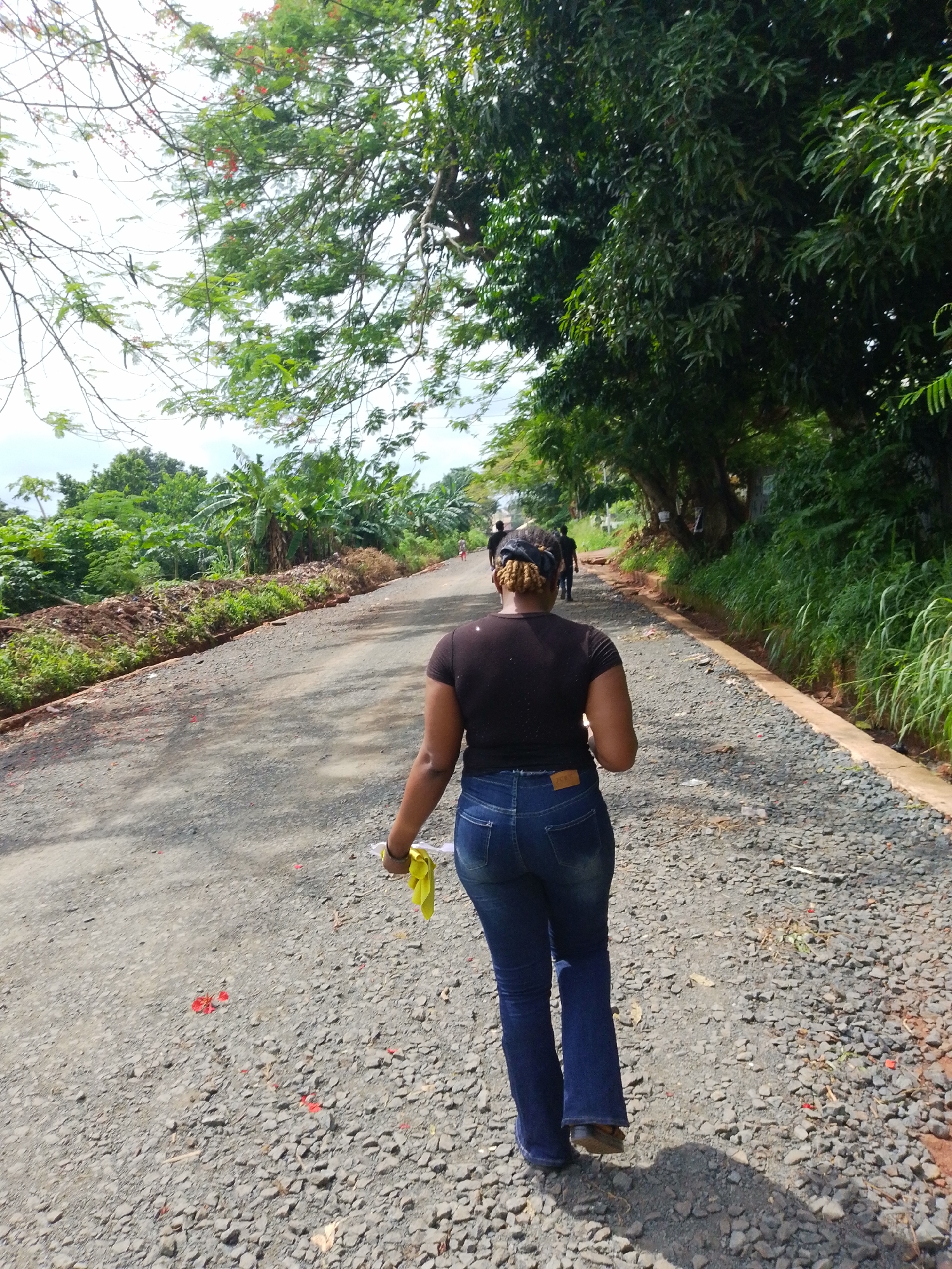 A low-angle, rear-view shot captures a woman walking away from the camera on a gravel road. She is wearing a black t-shirt and dark blue jeans, and a yellow cloth is clutched in her right hand. Her blonde-brown hair is pulled back in a bun with a black fabric band. The road is lined with dense green vegetation and trees on either side. In the distance, several other figures are walking further down the road. Red petals are scattered on the road surface. The scene appears to be outdoors in a rural or semi-rural setting within Nsukka, Nigeria.