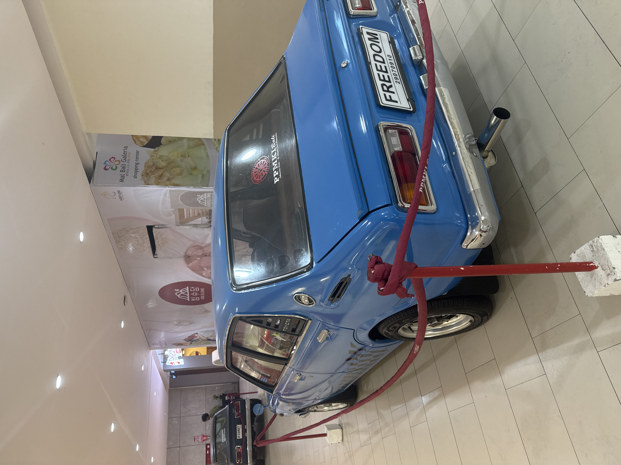 The image shows the back of a bright blue classic Toyota car, seemingly on display in a shopping mall. The car is surrounded by a red rope barrier held up by white concrete blocks. The license plate reads "FREEDOM 29070910." A sticker on the rear window reads "PPMKI Bali." In the background, there are advertisements for "Mal Bali Galeria" shopping center and a restaurant called "빙수당 BONG SU DANG." Another dark-colored vehicle, possibly a D'CORET car, is visible further back. The location appears to be in a shopping mall, indicated by the signage and tiled floor.