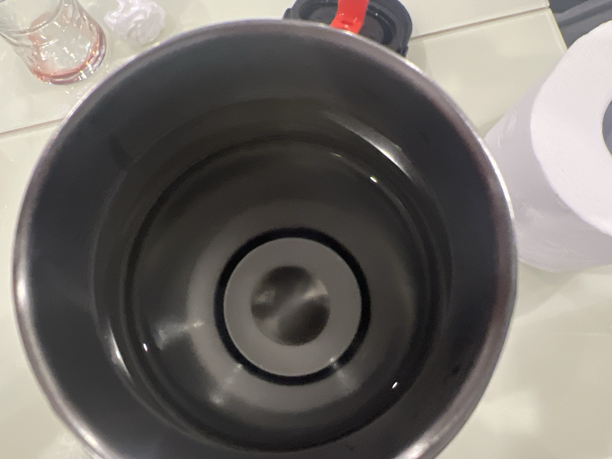 A close-up, overhead view of the interior of a dark-colored, metallic insulated tumbler filled with clear liquid. The tumbler has a stepped interior, with a central raised section. The tumbler is placed on a pale, tiled surface, possibly a countertop. To the upper right of the tumbler, a small, clear glass with a pinkish liquid is visible, along with a crumpled piece of white paper. A black lid with a red tab is partially visible to the right of the tumbler. A roll of white toilet paper is situated in the lower portion of the image. The scene appears to be indoors. There is no information or visual evidence to suggest a location in Podgumer, Bulgaria.