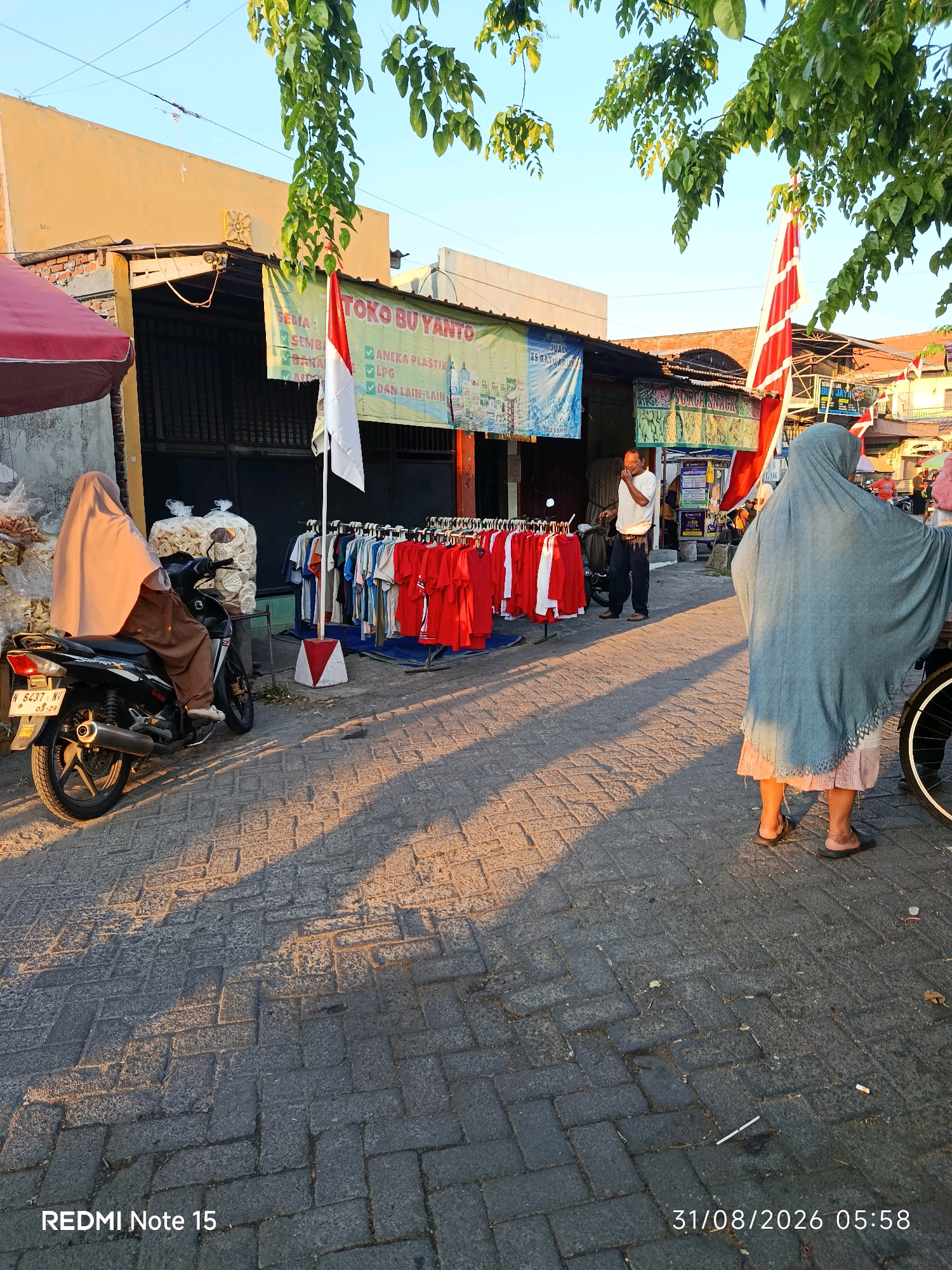In Keboansikep, Indonesia, an outdoor street market is captured during daylight. A woman, veiled in a light brown hijab and brown pants, sits on a black motorcycle, facing away from the camera. Beside her, a rack of red and white t-shirts is displayed for sale. In the background, a man in a white shirt and dark pants stands near a stall, appearing to converse. A large banner advertising "TOKO BU YANTO" is visible above the clothing rack, along with other signs indicating various goods. The street is paved with interlocking bricks, and shadows from trees and structures stretch across the scene. A second woman, veiled in a grey hijab and a light pink skirt, stands further down the street, facing away from the camera, near what appears to be a bicycle wheel. The sky is clear and blue.