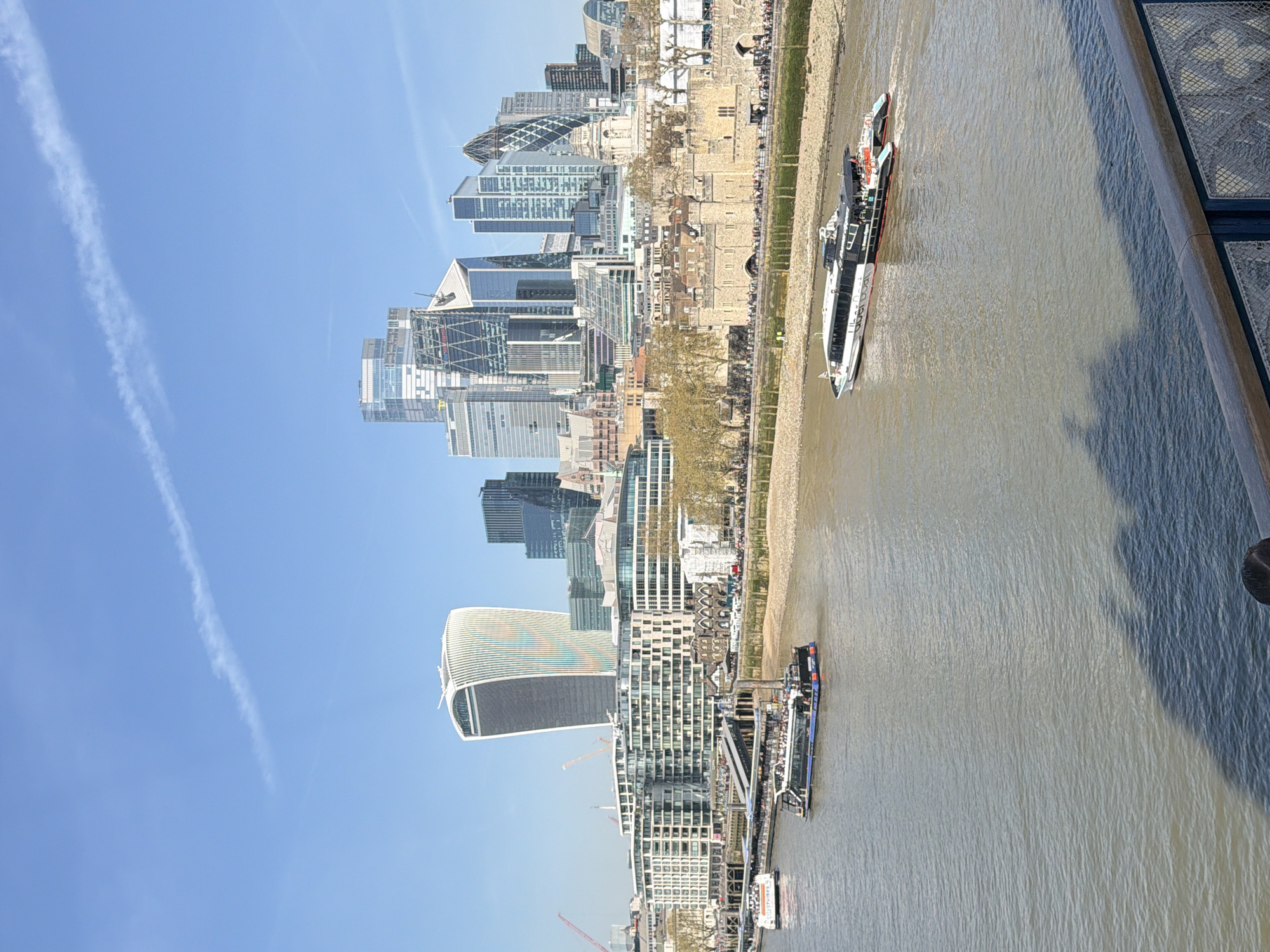 Here is a description of the image:
The image shows a cityscape in London, United Kingdom, likely taken from a high vantage point. The River Thames runs through the scene, with several boats visible on the water. Notable buildings include the Gherkin, the Cheesegrater, and the Walkie Talkie, all recognizable skyscrapers in the City of London.
Along the riverbank, there is a walkway populated by people, suggesting a tourist or pedestrian area. The Tower of London is visible near the river. The sky is a clear blue, with some contrails of planes.
The photo seems to be taken from a high bridge or building, with the edge of the structure visible in the foreground.