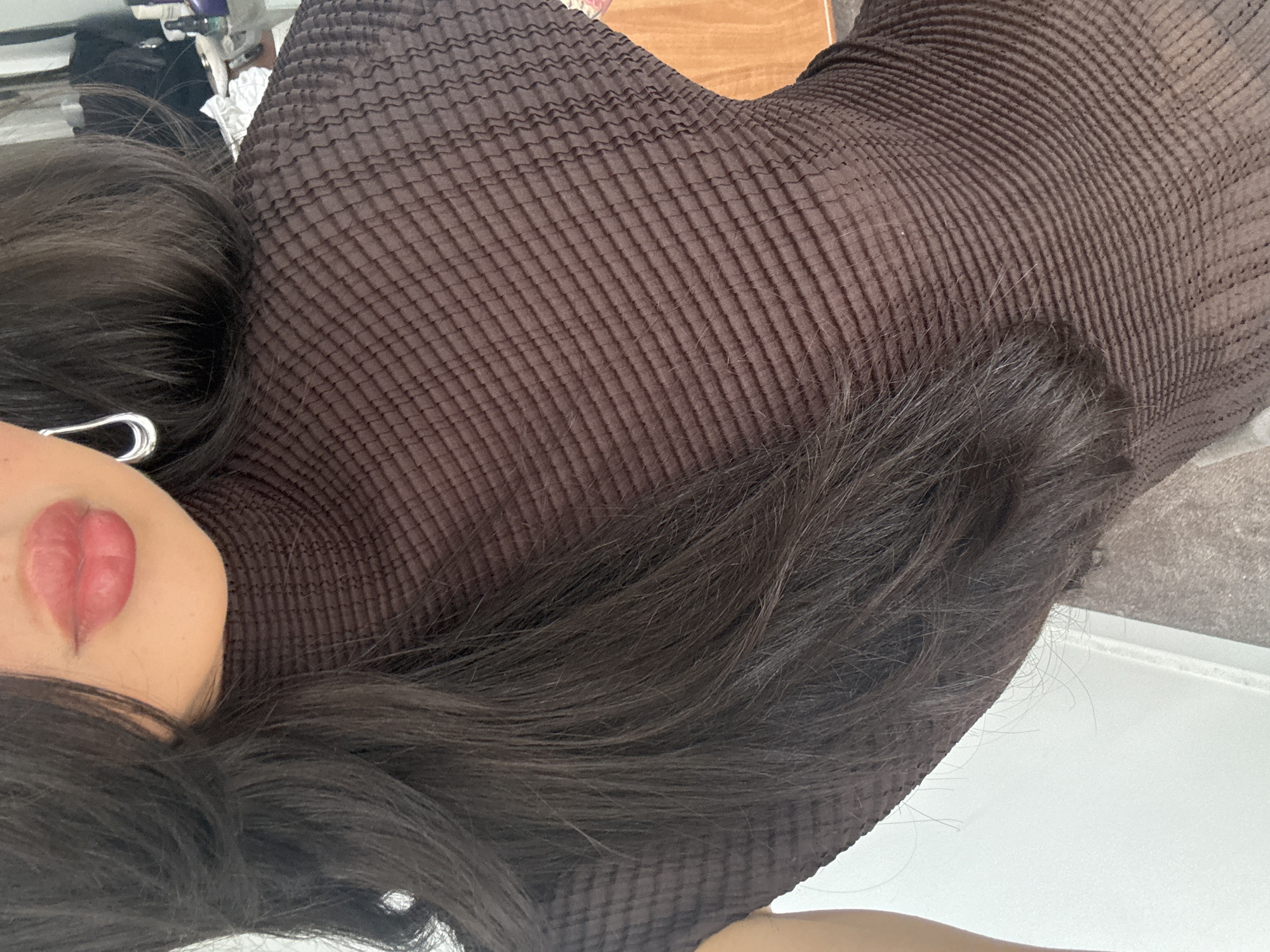A close-up, angled portrait features a person, likely female, with long, dark brown hair flowing across their shoulders and chest. The subject is wearing a dark brown garment, possibly a high-neck top or dress, which exhibits a distinctive finely pleated or ribbed texture. Part of the face is visible, including the lips, which are coated with a subtle pink lipstick, and a hint of the chin. A silver, C-shaped earring is visible near the person's left ear.
The image is composed as a close-up, with the background largely out of focus. This blurred setting suggests an indoor environment, possibly a bedroom or living space. Visible elements in the background include a wooden surface, such as a headboard or table, and lighter areas that could be bedding or walls. Vaguely discernible objects, possibly toiletries or other household items, are present in the top left corner of the blurred background.
The person appears to be posing for the photograph, with no specific activity taking place other than being captured in a static pose. The expression conveyed by the visible lips is neutral or relaxed. There are no overt location cues within the image to confirm the London, United Kingdom context, nor are there any indications of the time of day, weather, or visible text. The intricate texture of the clothing and the flowing dark hair are notable visual details.