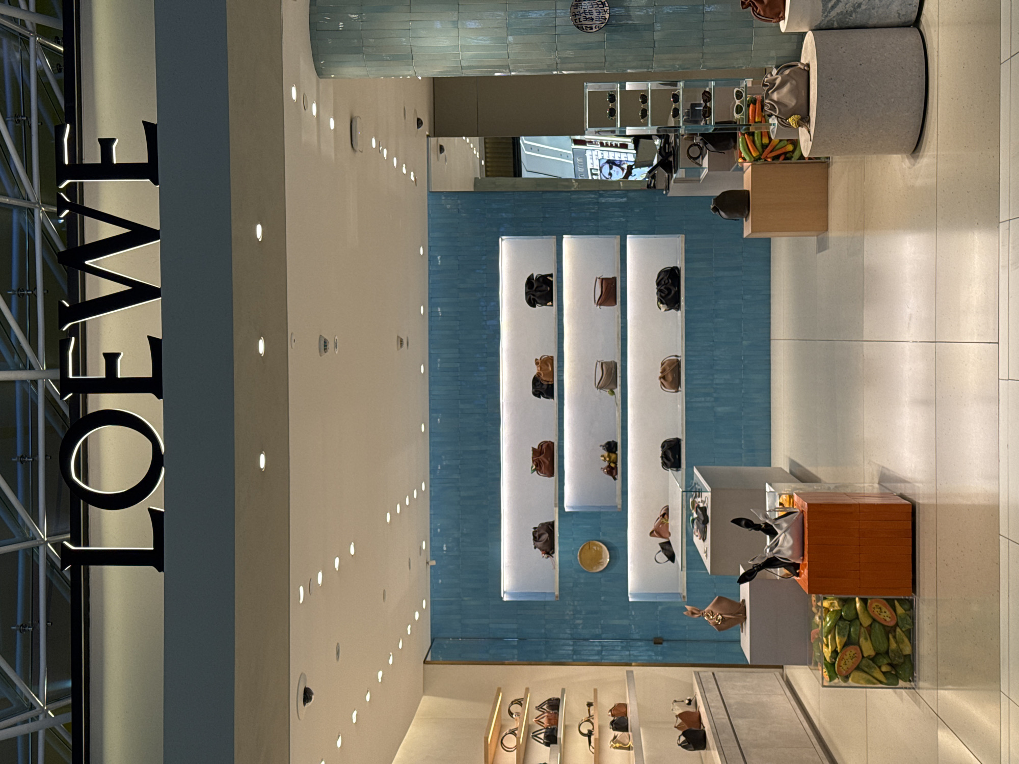 The image depicts the interior of a luxury retail store, likely a handbag or fashion boutique, in Nong Prue Subdistrict, Thailand. The store features modern, minimalist displays with white shelving and light-colored walls. A prominent "LOEWE" logo is visible on the left side of the image, indicating the brand.
Various handbags are displayed on shelves. One display features a clear cube filled with sliced fruits, possibly as a decorative element. Another display shows a cylindrical concrete pedestal with a handbag on top. The store lighting is soft and ambient, suggesting it could be evening or the store is designed with low lighting. The overall aesthetic is clean, sophisticated, and upscale. No people are visible in the image, and the scene appears to be a quiet moment within the store. The tiled blue wall in the background adds a touch of color and texture to the otherwise neutral palette.