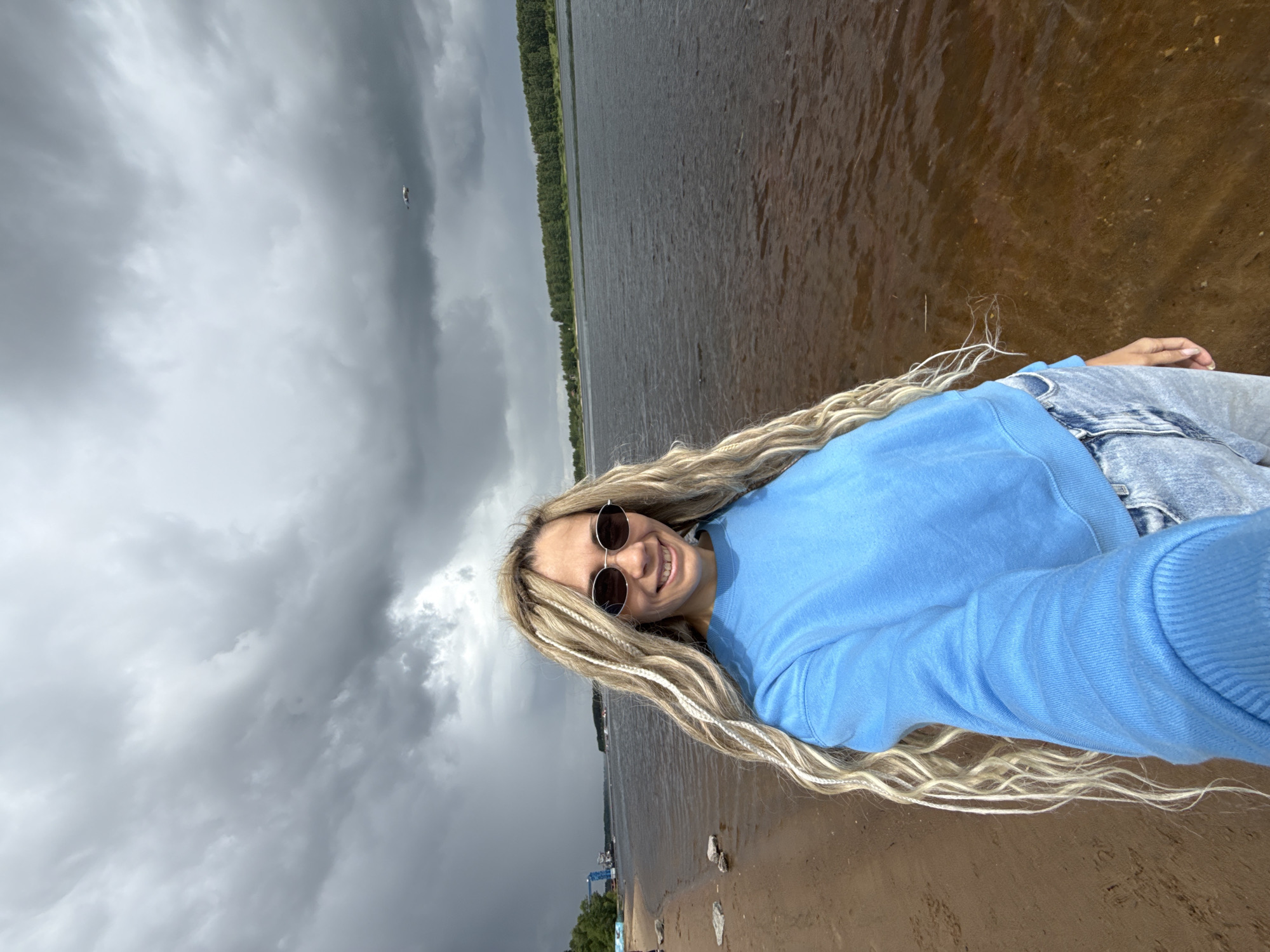 A selfie taken at Rusanovo, Russia, shows a woman with blonde braids, wearing a blue sweatshirt and round sunglasses, standing near a body of water. The sky is overcast with dark clouds. In the background, trees line the opposite shore. The foreground consists of sand and shallow water.