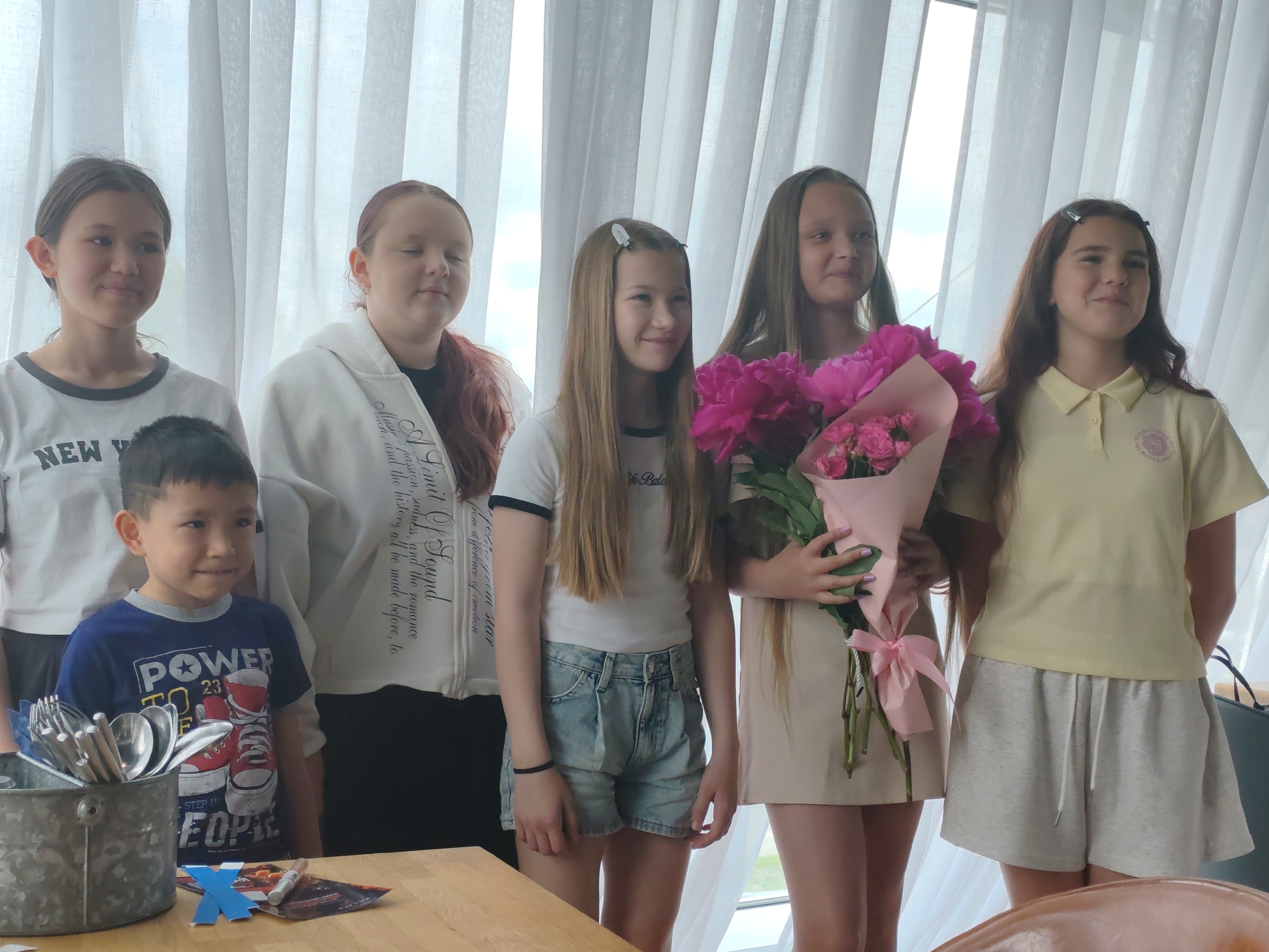 A group of six children, five girls and one boy, are posed in front of a white, sheer curtain. The girl on the far left has dark hair pulled back and is wearing a white t-shirt with "NEW YORK" printed on it. Next to her is a young boy wearing a blue t-shirt with a graphic design and the words "POWER TO 23." He is smiling slightly. To the boy's right is a girl with reddish-brown hair, wearing a white hooded sweatshirt with text and designs. She has her eyes closed. The next girl, with long, straight blonde hair, wears a white t-shirt and denim shorts. She is holding a bouquet of pink peonies and smaller pink flowers. Next to her is another girl with long blonde hair, wearing a light yellow polo shirt. The girl on the far right has long brown hair and is wearing a pale yellow t-shirt. All the children appear to be standing on a wooden surface, and in the foreground, a metal bucket filled with silverware, two blue paper cutouts, and a pen are visible. The background suggests an indoor setting with a window behind the curtain. The overall scene appears to be a staged photograph, possibly a celebration or a school event. No specific landmarks of Amursk, Russia, are identifiable in the image.
