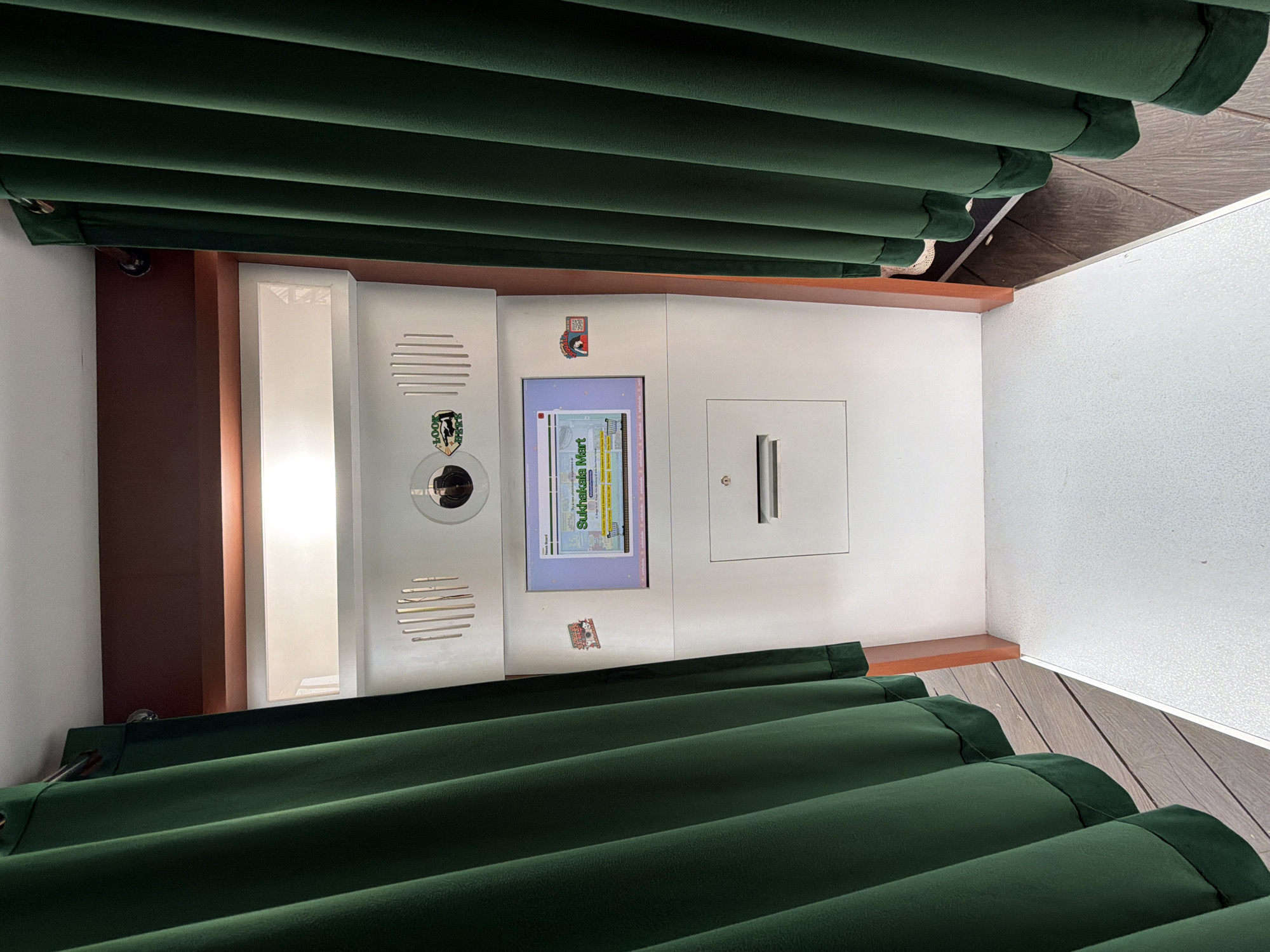The image depicts an ATM-like machine in what appears to be an indoor setting. The machine has a screen displaying the words "Sukhalakela Mart". There are green curtains partially obscuring the top and bottom of the machine. The environment includes a white wall and wood-paneled accents. Based on the text on the machine, it is located in Candikuning, Indonesia.