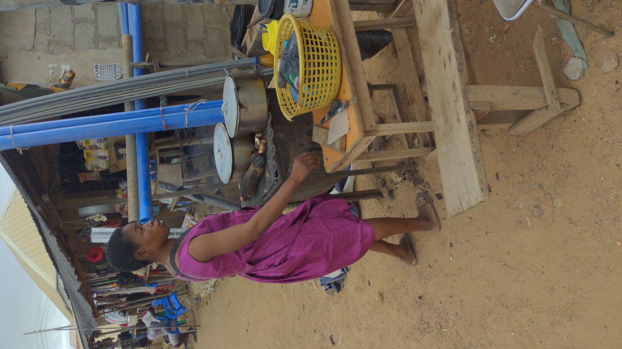 A woman in a purple dress stands near a cooking area on a sandy ground in what appears to be Masaka, Nigeria. She faces towards the right of the frame, towards several pots sitting atop a charcoal grill. The pots are covered with lids and presumably cooking food. Beside the pots is a yellow plastic basket. The cooking area appears to be near a building with construction supplies, including blue plastic pipes. A partially visible poster is affixed to the building. A wooden structure is in the foreground on the right. The sky is overcast.