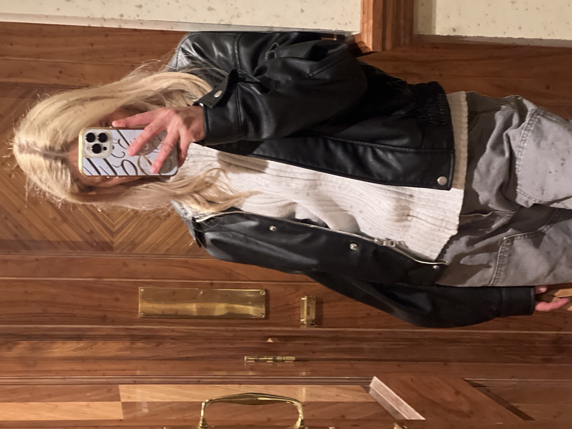 A blonde woman takes a selfie with a phone in a reflective doorway in London, United Kingdom. She wears a black leather jacket, a white sweater, and gray pants. The door is made of wood, with brass hardware including a handle, mail slot, and hinges. The doorway features decorative wooden molding.