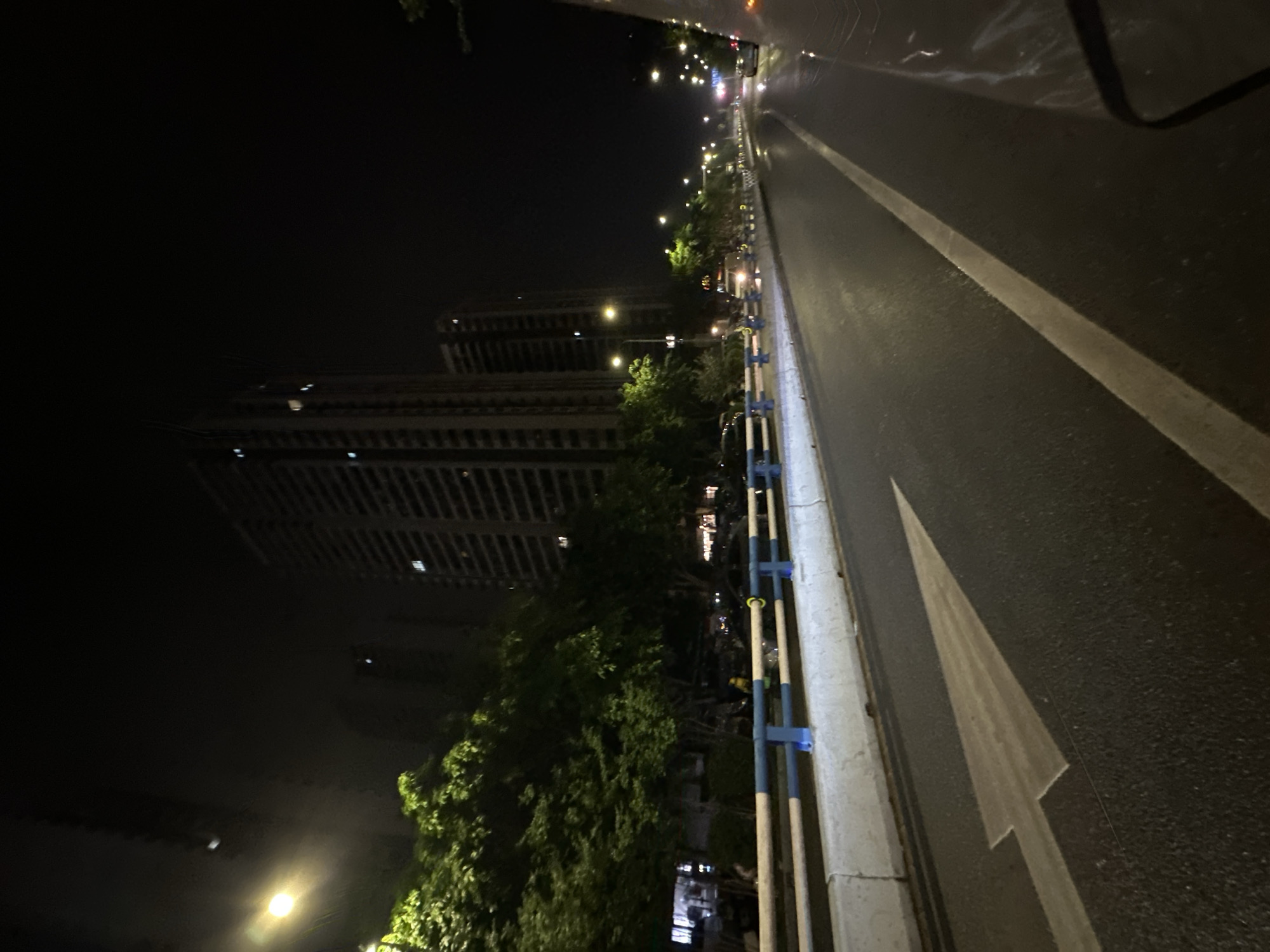 The image depicts a nighttime urban scene captured from a moving vehicle on a multi-lane road in the Chongqing High-tech Zone, China. The asphalt road surface is wet and highly reflective, suggesting recent rain.
In the foreground, a large, white, forward-pointing arrow is painted on the pavement of the left lane, indicating the permissible direction of travel. A solid white line runs along the right edge of the current lane, separating it from an adjacent lane or shoulder. To the left, a concrete median barrier topped with a blue and white railing runs parallel to the road.
Beyond this barrier, a dense line of green trees extends along the roadside. Behind the trees, multiple tall, multi-story buildings with numerous illuminated windows rise prominently against the dark night sky, indicating a dense urban or residential area. Another line of similar buildings is partially visible further down the road on the right.
The scene is illuminated by numerous streetlights with a warm, yellowish glow, casting significant reflections on the wet road surface. These lights recede into the distance along both sides of the road. Faint vehicle lights are visible further ahead, suggesting other traffic. A portion of the camera's originating vehicle, possibly its hood or side panel, is visible in the lower right corner of the frame. No clear individuals are discernible within the scene; however, a yellow, indistinct object is present behind the left barrier amidst the trees. The sky is dark and featureless.