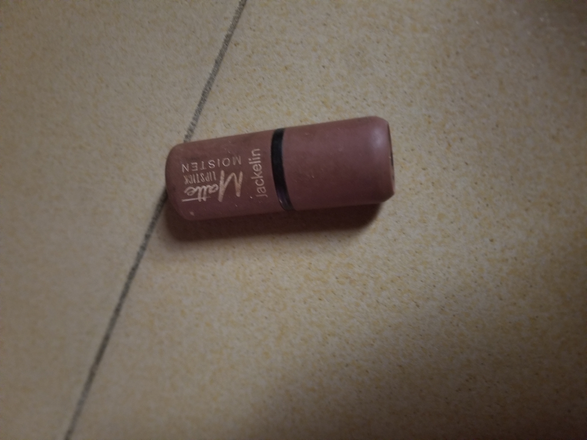 A single tube of matte lipstick, a shade of dusty rose, lies horizontally on a speckled tan floor. The lipstick is partially opened, revealing the tip. White text on the side of the tube reads "Matte Lipstick Moisten Jackelin". The floor appears to be tiled, with a thin, dark line running diagonally across the upper left portion of the image. No people or other objects are visible. The lighting suggests an indoor environment. The image is taken from a slightly elevated angle looking down. The location is Jauro Yinu, Nigeria.