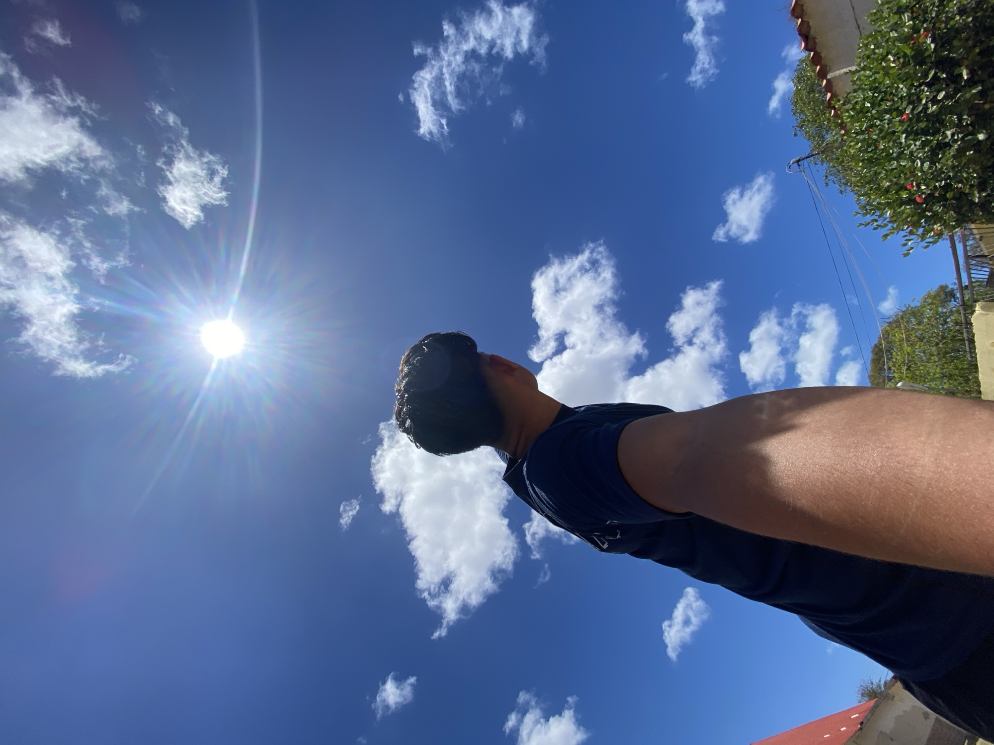 A low-angle shot of a man looking up at the sky in Juan Griego, Venezuela. The sun is visible in the upper left, surrounded by a blue sky with scattered white clouds. Part of a building and green foliage are in the upper right. The man is wearing a dark blue shirt.