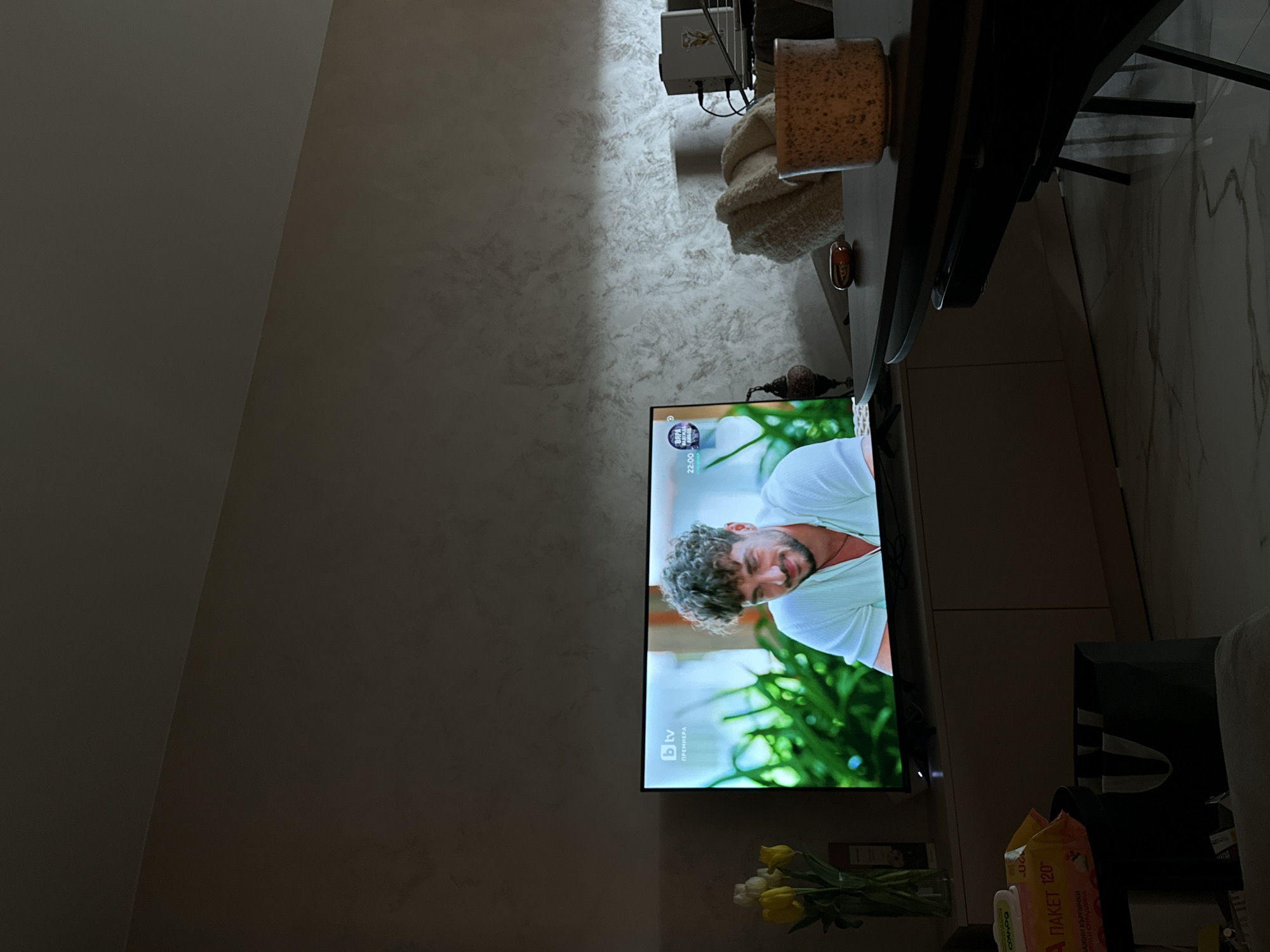 The image captures an interior residential scene, likely a living or dining area, illuminated by dim ambient light, situated in Sofia, Bulgaria. A flat-screen television, positioned centrally, displays a program from bTV, a Bulgarian channel, featuring a smiling male with dark, curly hair wearing a light-colored polo shirt, set against a blurred green, possibly outdoor, background. On-screen text indicates "bTV ПРЕМИЕРА" (bTV Premiere) and a time of "22:00". The television rests on a light-colored console cabinet with two doors. To the left of the console, a clear glass vase contains yellow and white tulips. Adjacent to the vase, a package of wet wipes bearing Cyrillic text ("ПАКЕТ 120") is visible on a lower surface, with a dark bag beneath it. In the mid-ground right, a dark dining table holds a textured, light brown ceramic mug and a small, reflective brown object. Further right and in the background, a light-colored, textured blanket is draped over a piece of furniture, behind which electronic devices, including a white box and a lamp, are visible. The walls feature a textured, mottled finish in shades of grey and beige, and the floor is composed of light-colored, veined marble or tile.